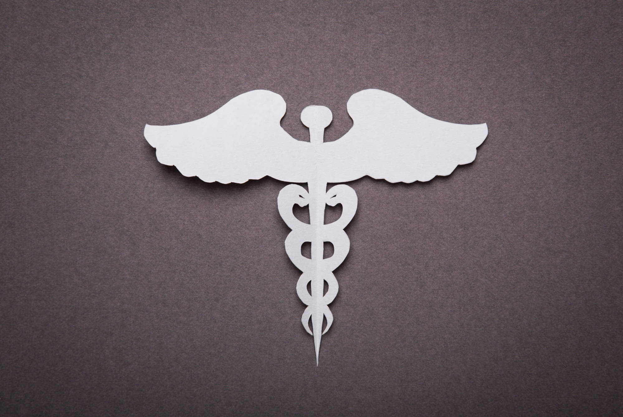 medical motivation 桌布 壁紙 — 一個白色紙剪成的醫療雙蛇杖標誌，置於有紋理的深灰色表面中央。