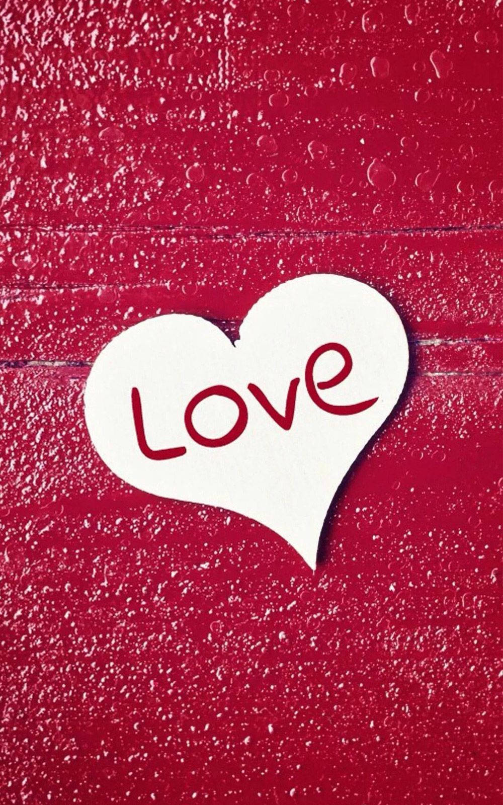 Love Phone 桌布 壁紙 — 一個白色紙愛心，上面有紅色草書 'Love'，放在有水滴的紋理紅色表面上。