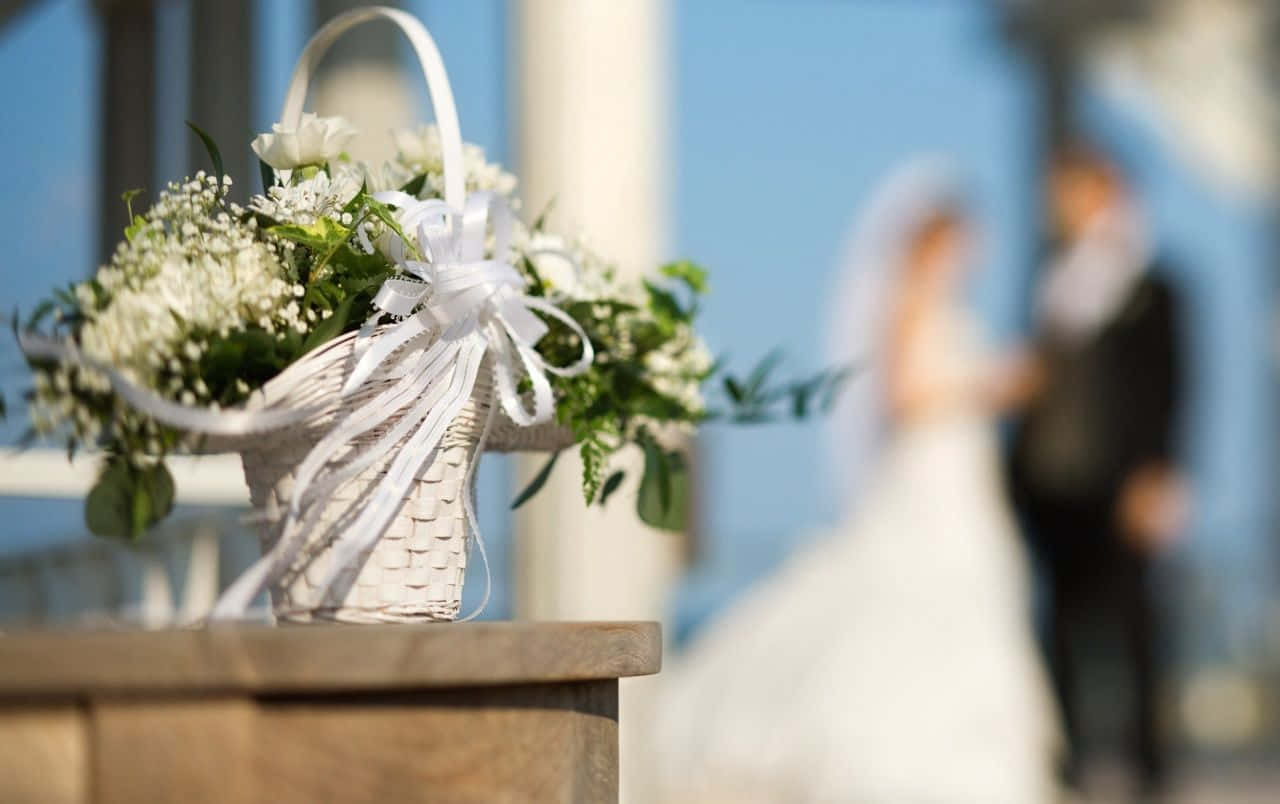 Wedding Flowers 桌布 壁紙 — 一個白色編織籃子裝滿滿天星和白色鮮花，放在木架上，背景有一對模糊的新郎新娘。