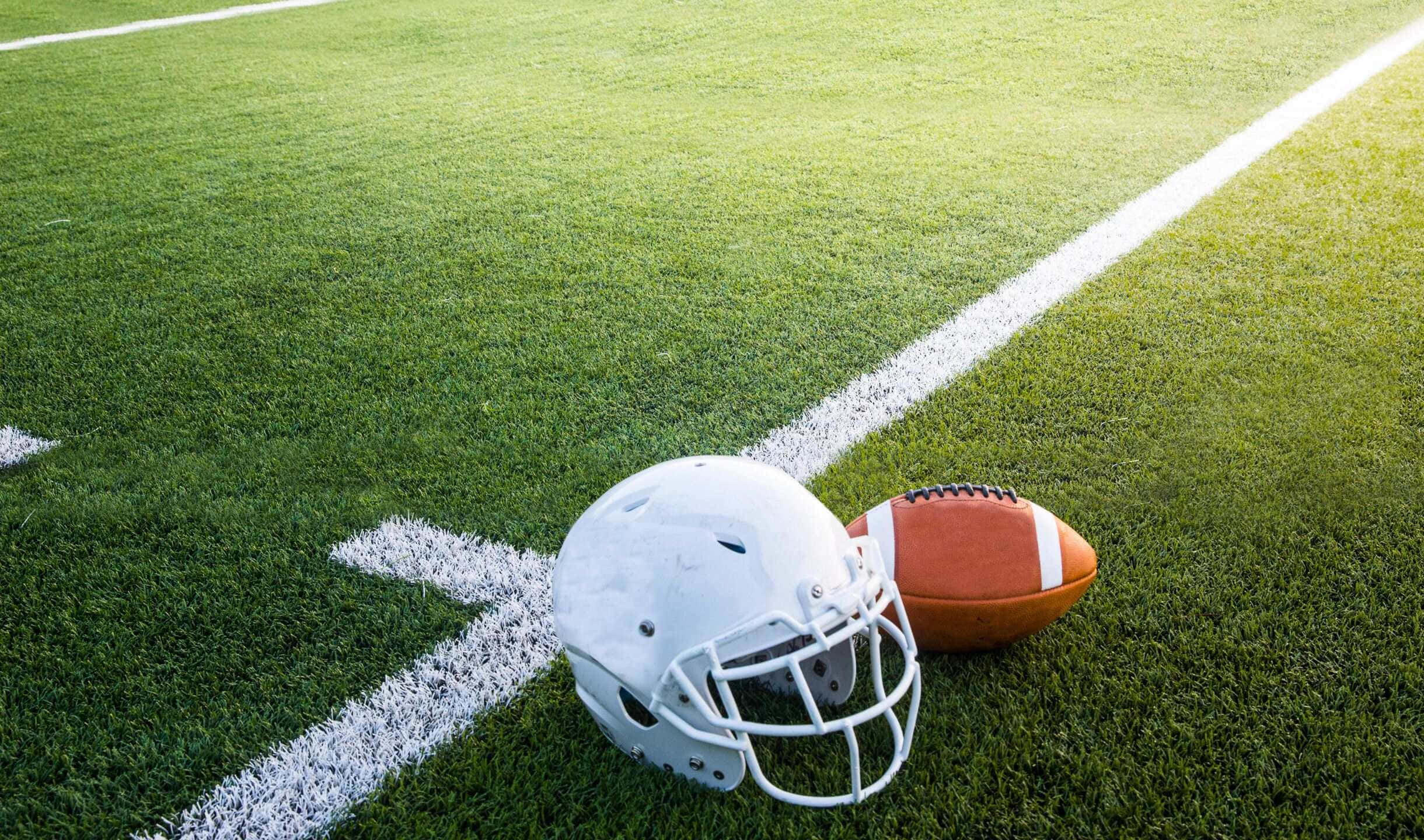 運動 桌布 壁紙 — 一個白色美式足球頭盔和一個棕色皮球放在鮮綠色草地上，草地上有清晰的白色線條，在明亮的陽光下。