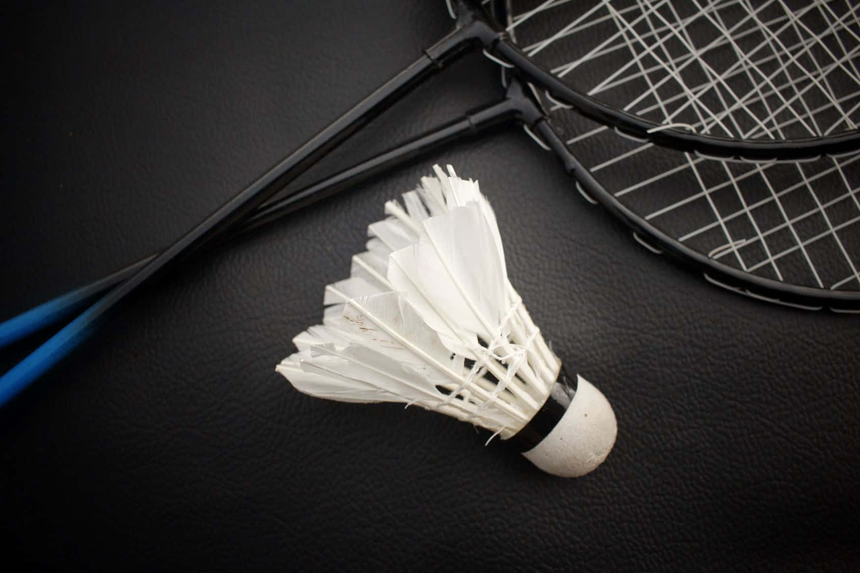 HD Badminton 桌布 壁紙 — 一個白色羽毛球和兩個黑色羽毛球拍放在有紋理的黑色皮革表面上。