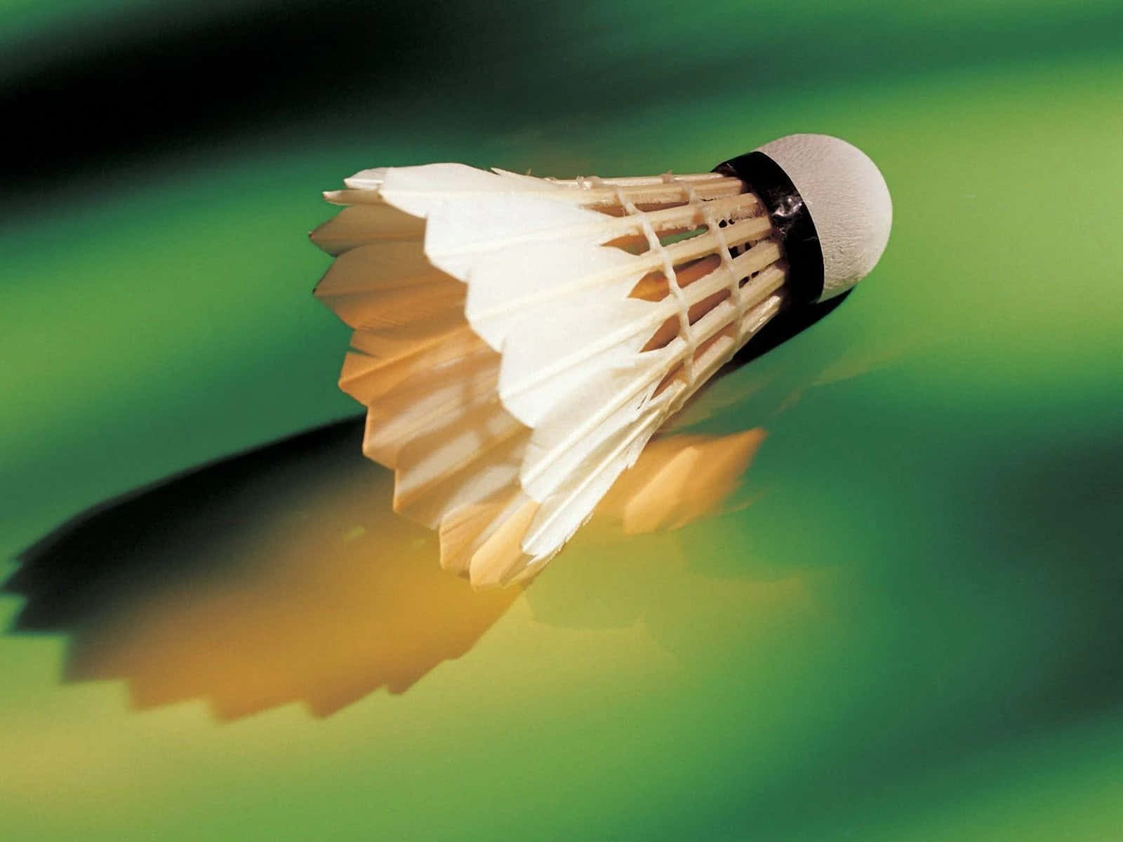 Best Badminton 桌布 壁紙 — 一個白色羽毛球在半空中飛行，在充滿活力的綠色漸變背景上投下清晰的陰影。