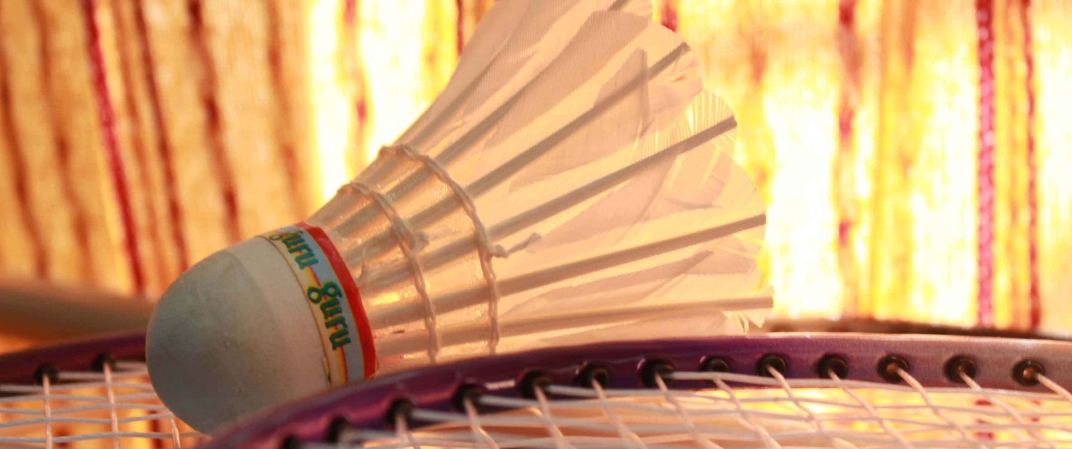 3440x1440p Badminton 桌布 壁紙 — 一個白色羽毛球的特寫，放在紫色羽毛球拍上，背景是溫暖的金色條紋。