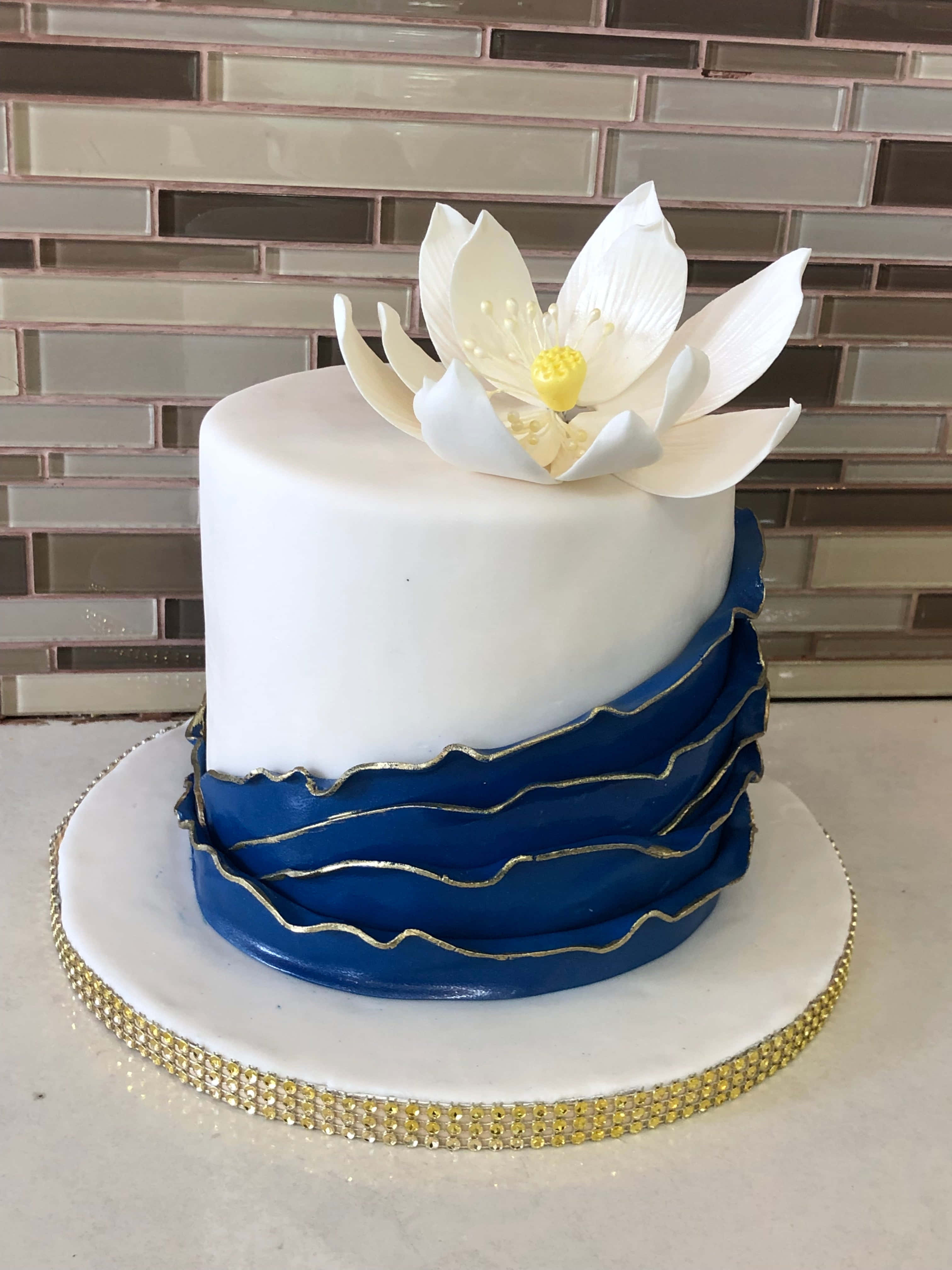 Cake Fondant 桌布 壁紙 — 一個白色翻糖蛋糕，帶有藍色皺褶層和白色蓮花裝飾，放在金邊大理石板上，背景是瓷磚牆。
