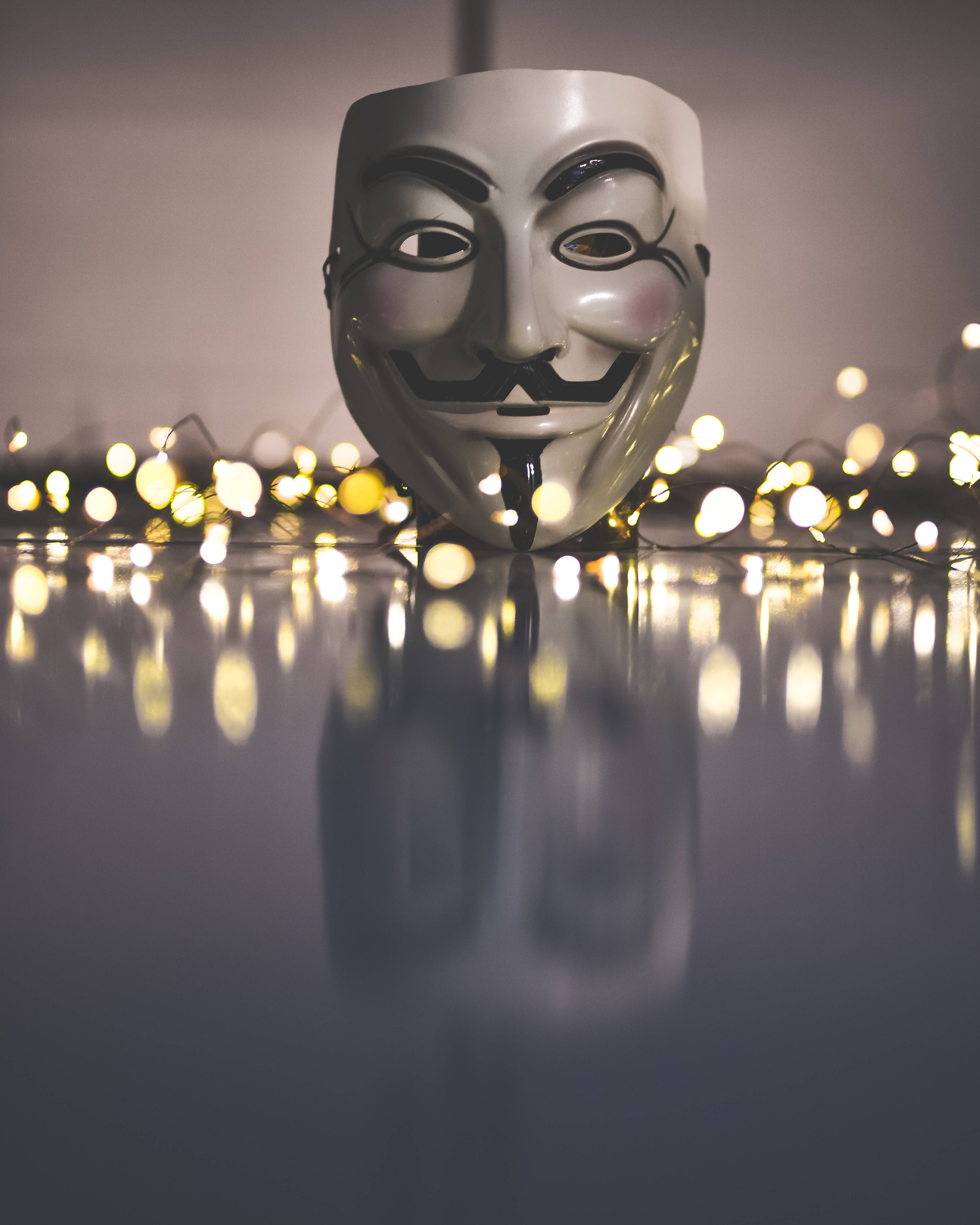 Bokeh 桌布 壁紙 — 一個白色蓋伊·福克斯面具放在反光表面上，周圍環繞著發光的燈串，營造出柔和的散景效果。