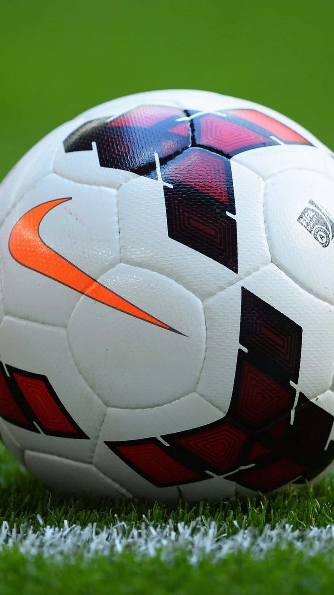 Soccer iPhone 桌布 壁紙 — 一個白色足球的特寫，帶有紅色、黑色和橙色的點綴，放在充滿活力的綠色草地上，靠近一條白色邊界線。
