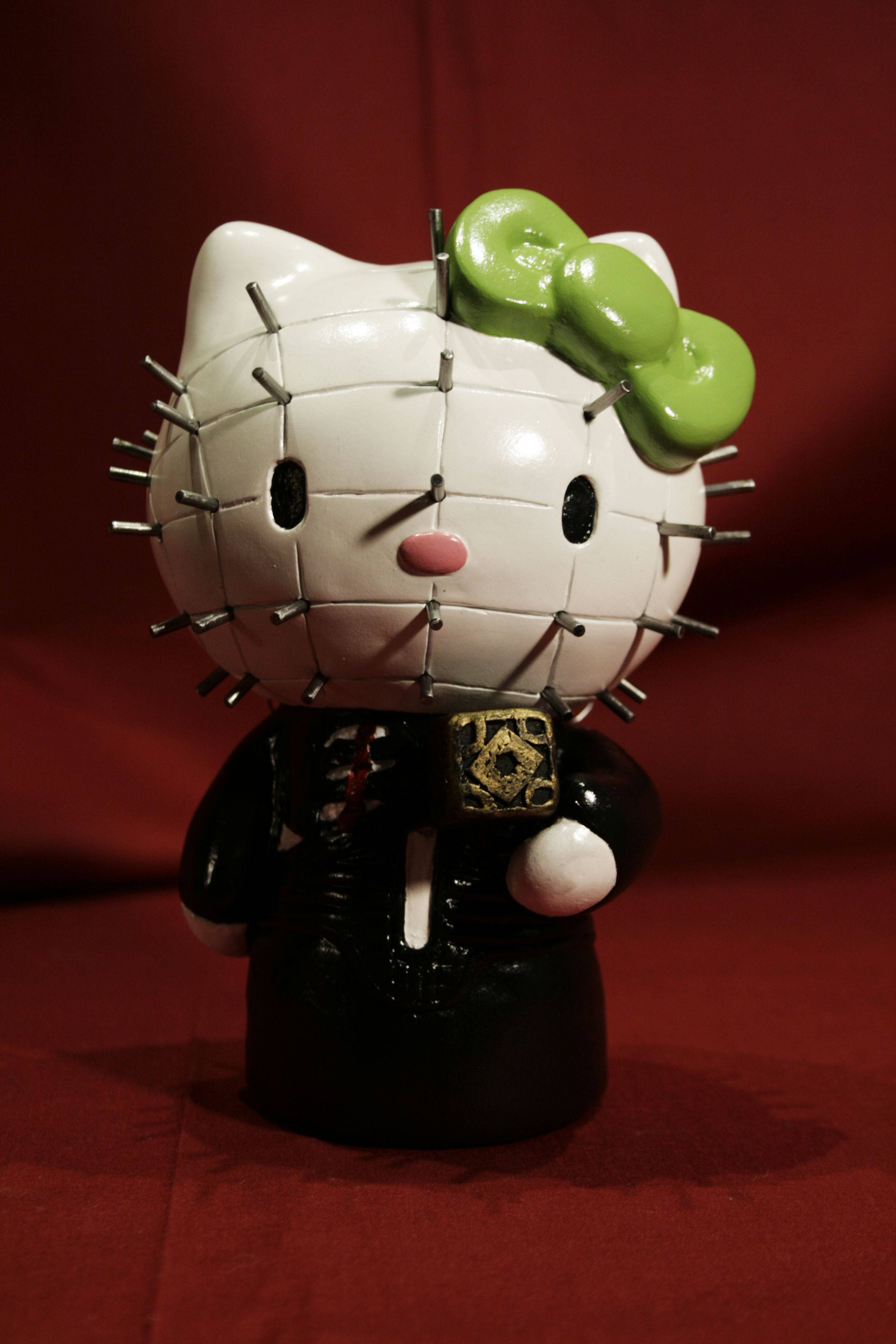 Hello Kitty Halloween 桌布 壁紙 — 一個白色陶瓷Hello Kitty公仔，頭上和身上有綠色蝴蝶結和金屬針，站在深紅色背景前。