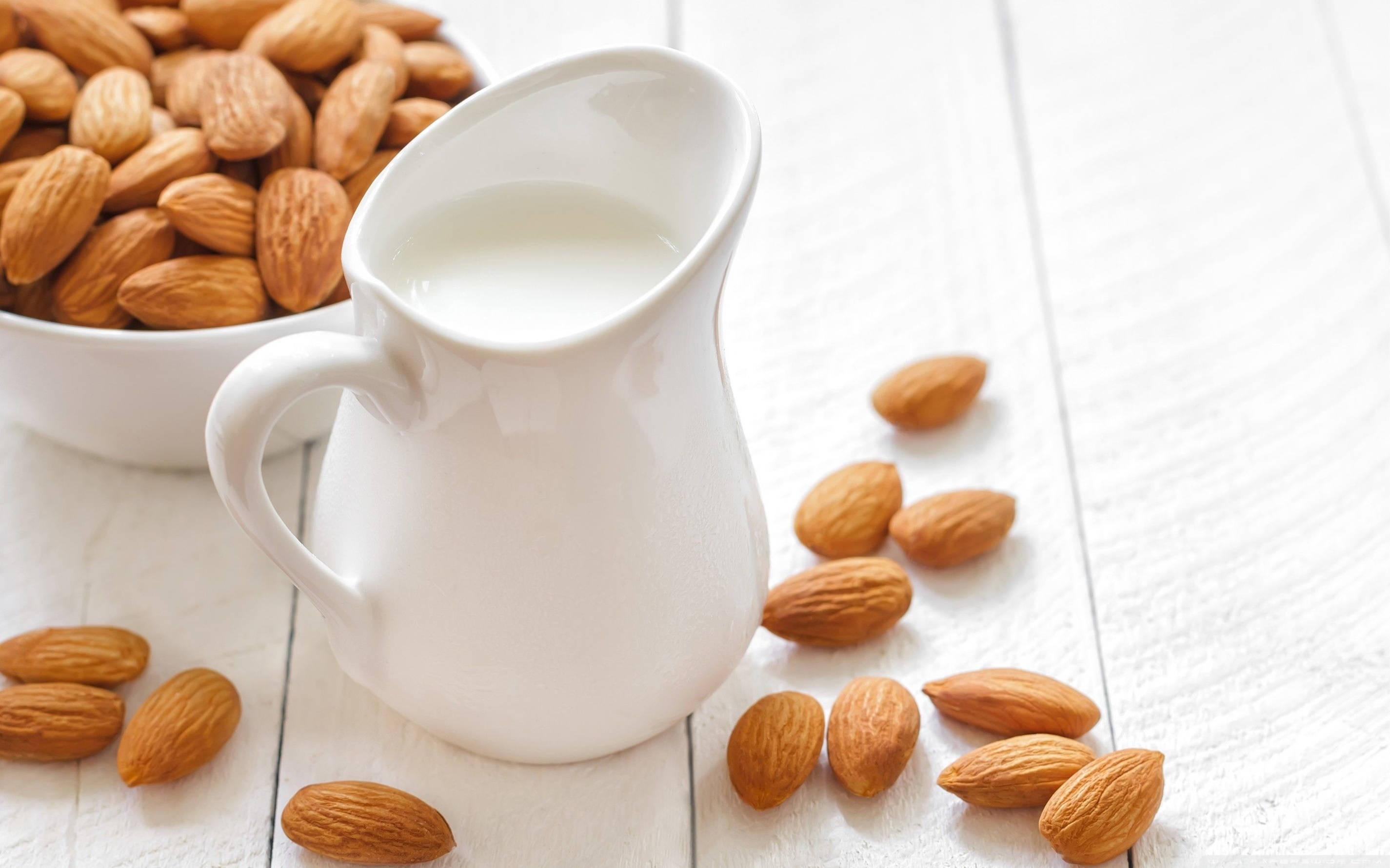 Milk 桌布 壁紙 — 一個白色陶瓷壺裝滿了奶油杏仁奶，旁邊有一個碗和散落的杏仁，放在明亮的白色木質表面上。