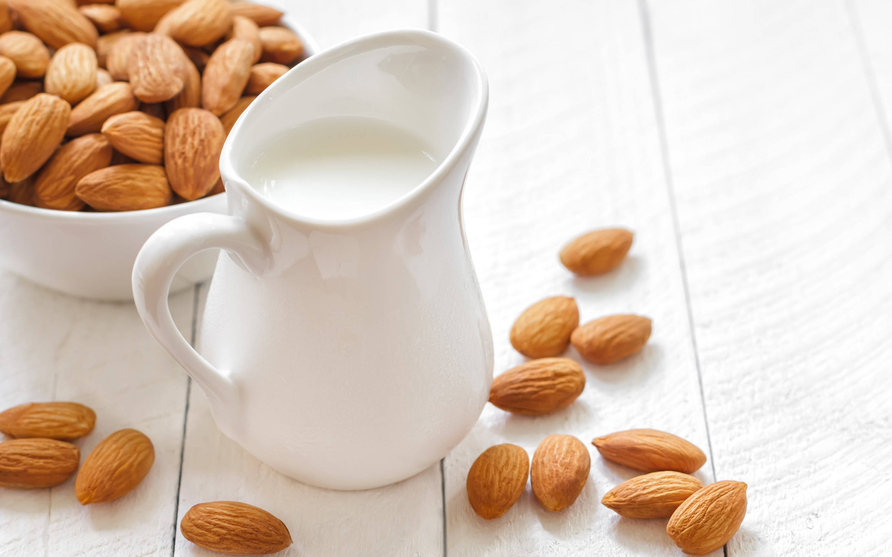Milk 桌布 壁紙 — 一個白色陶瓷壺裝滿了奶油杏仁奶，旁邊放著一個碗和散落的杏仁，在明亮的白色木質表面上。