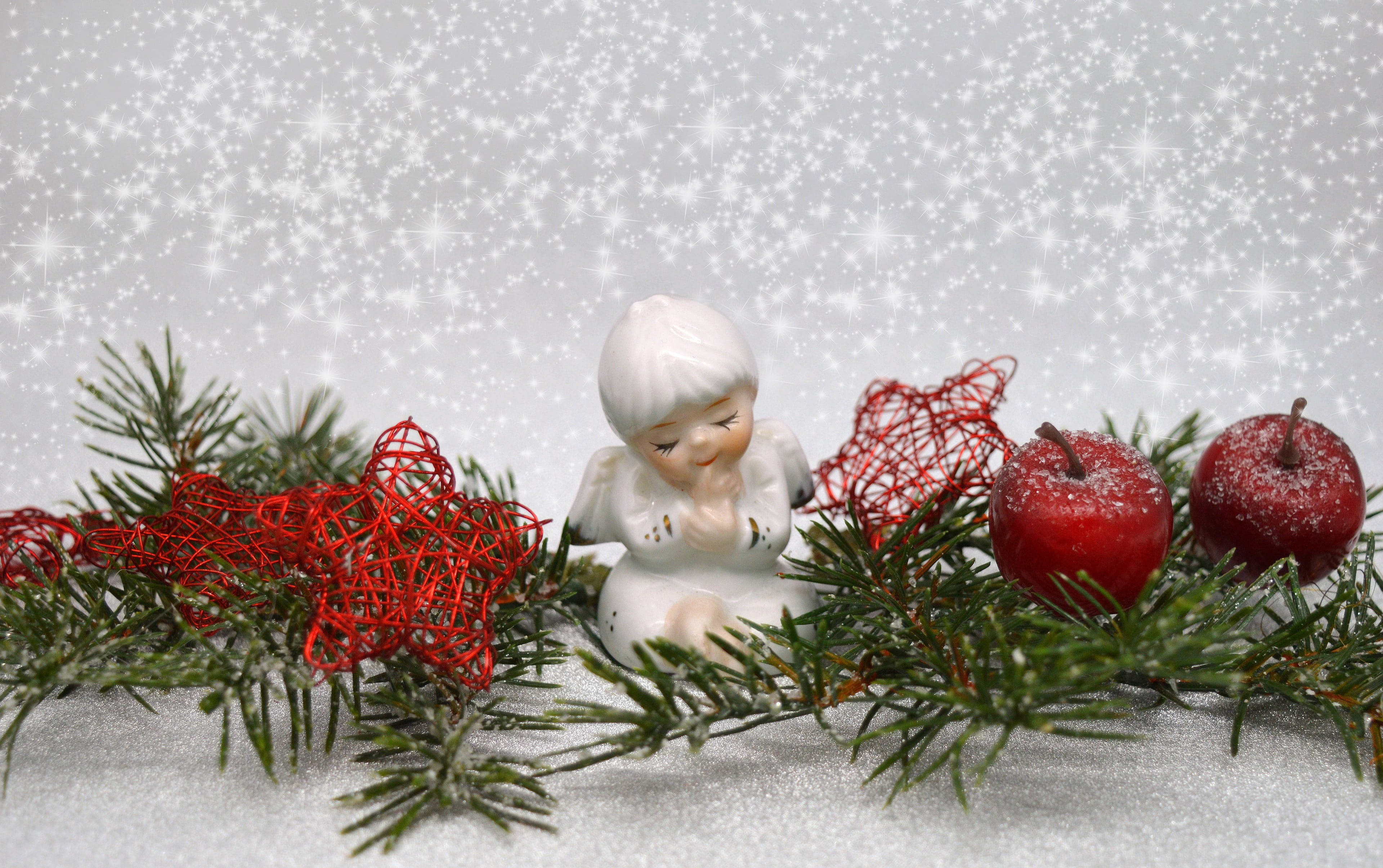 angels 桌布 壁紙 — 一個白色陶瓷天使雕像坐在白雪覆蓋的松樹枝、紅色鐵絲心和結霜的蘋果之間，背景是閃閃發光的冬日景色。