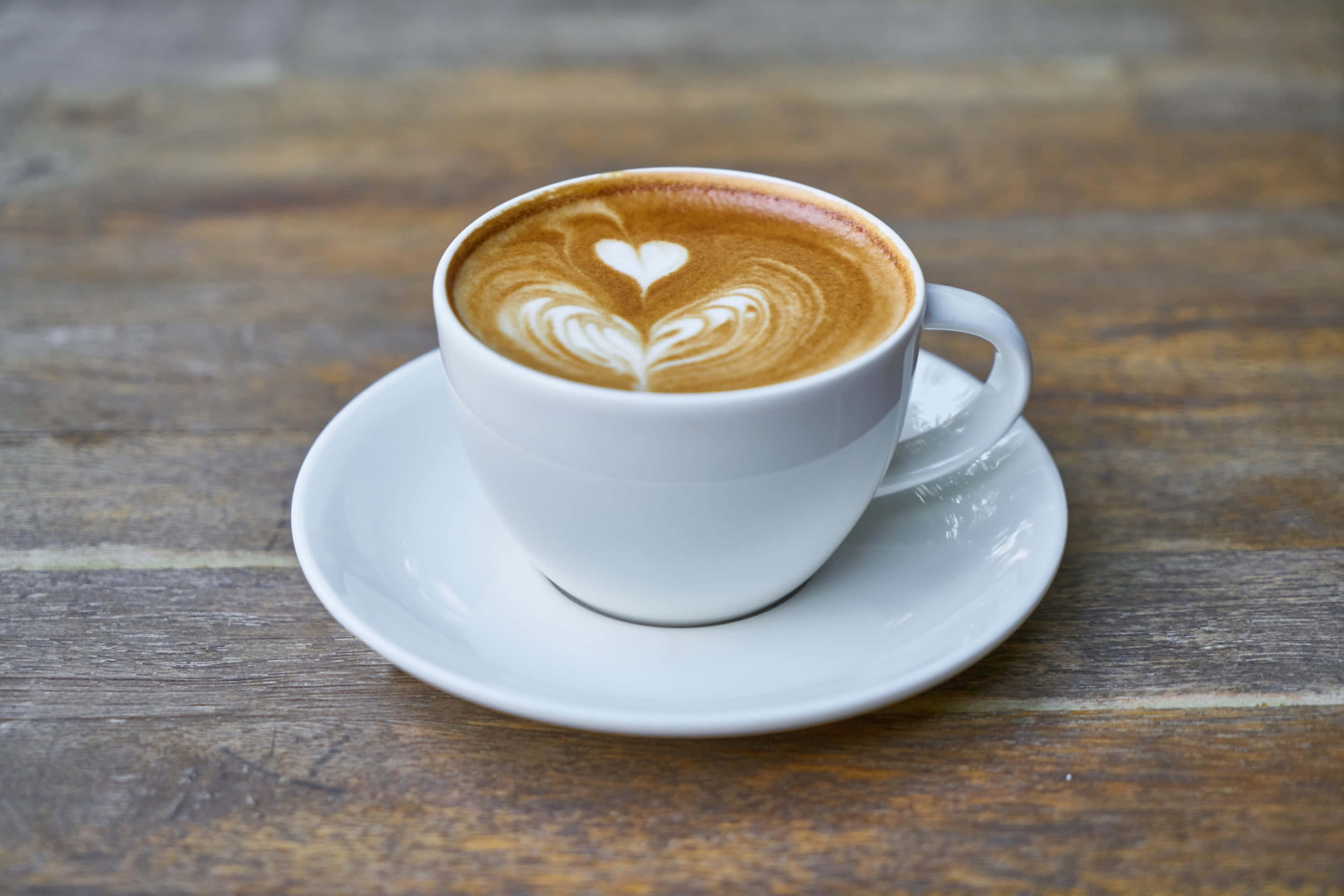 Coffee Cup 桌布 壁紙 — 一個白色陶瓷杯，上面有心形拉花藝術，放在一個樸實的木桌上的碟子上。