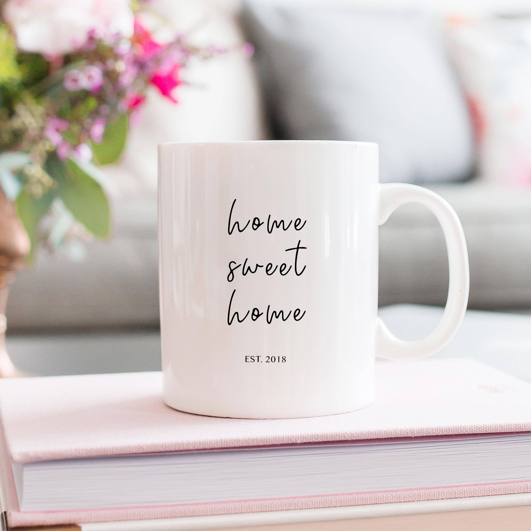 Home 桌布 壁紙 — 一個白色陶瓷杯，上面有黑色字體的 'home sweet home EST. 2018'，放在粉紅色書本上，旁邊有花卉擺設，置於舒適的客廳桌上。