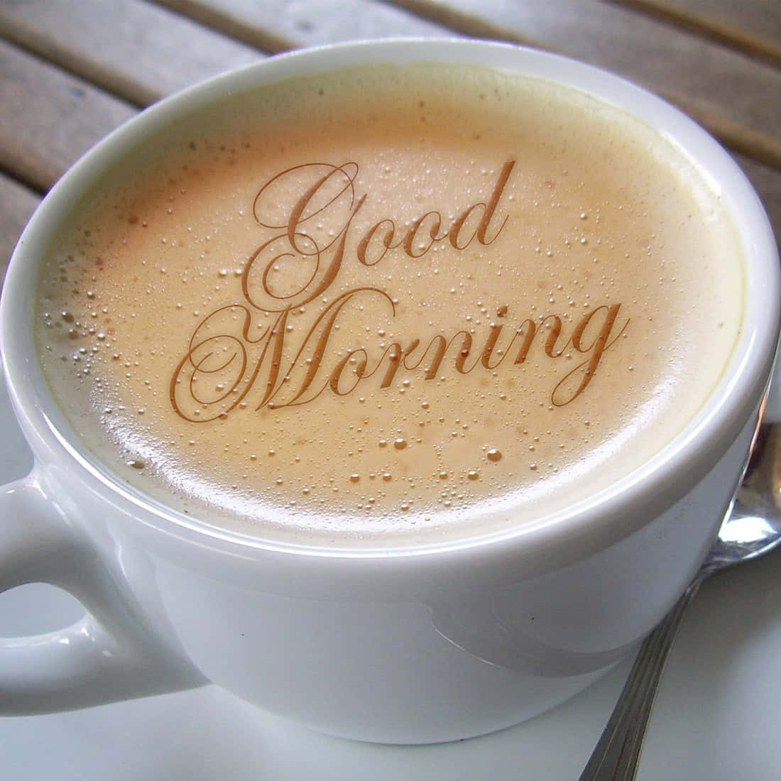 Morning Coffee 桌布 壁紙 — 一個白色陶瓷杯，盛滿泡沫咖啡，泡沫上優雅地刻有草書「Good Morning」，放在木桌上，旁邊有一隻湯匙。