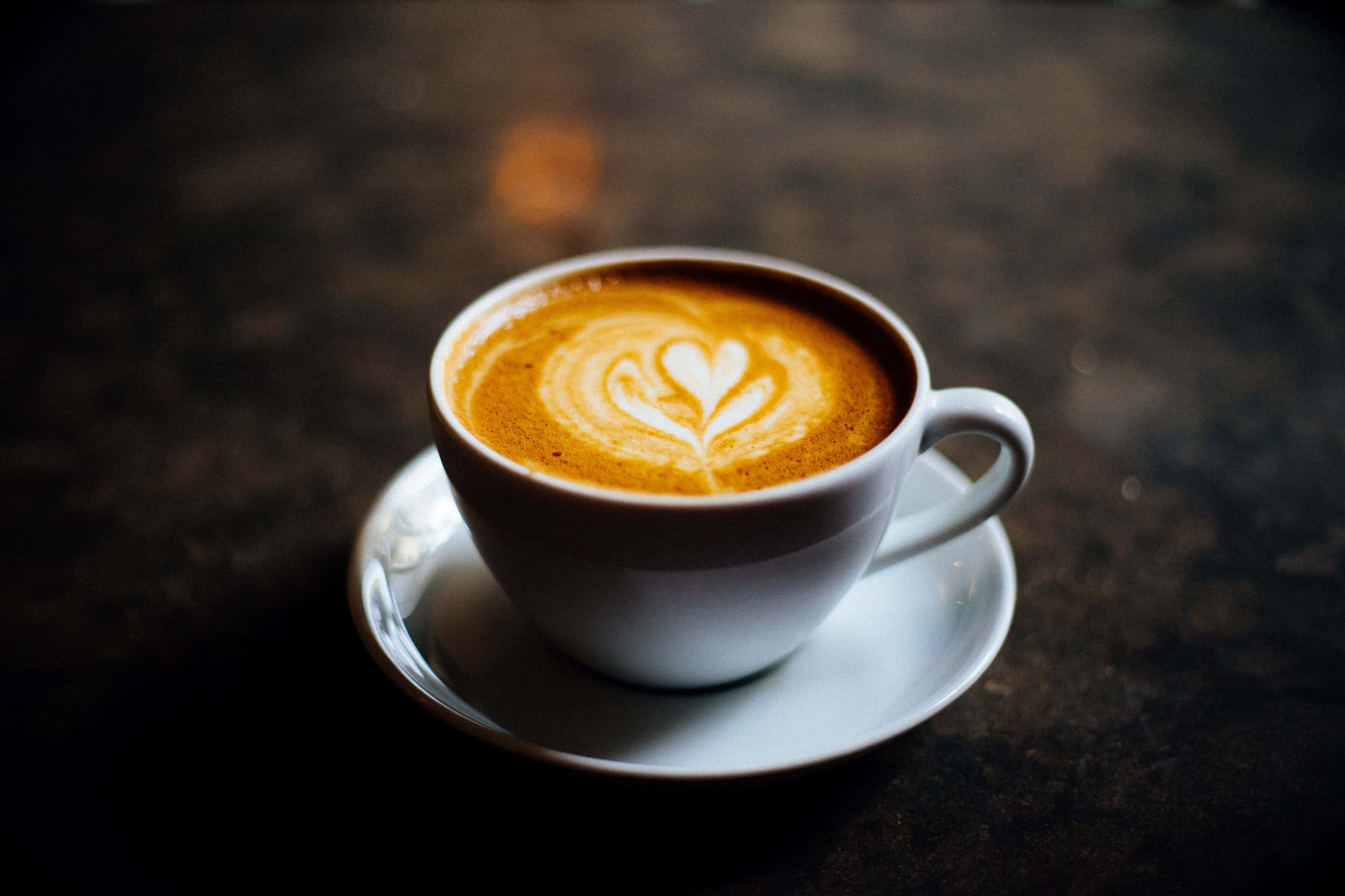 Coffee Cup 桌布 壁紙 — 一個白色陶瓷杯，盛載著帶有心形圖案的拉花咖啡，放在碟子上，背景為深色紋理。