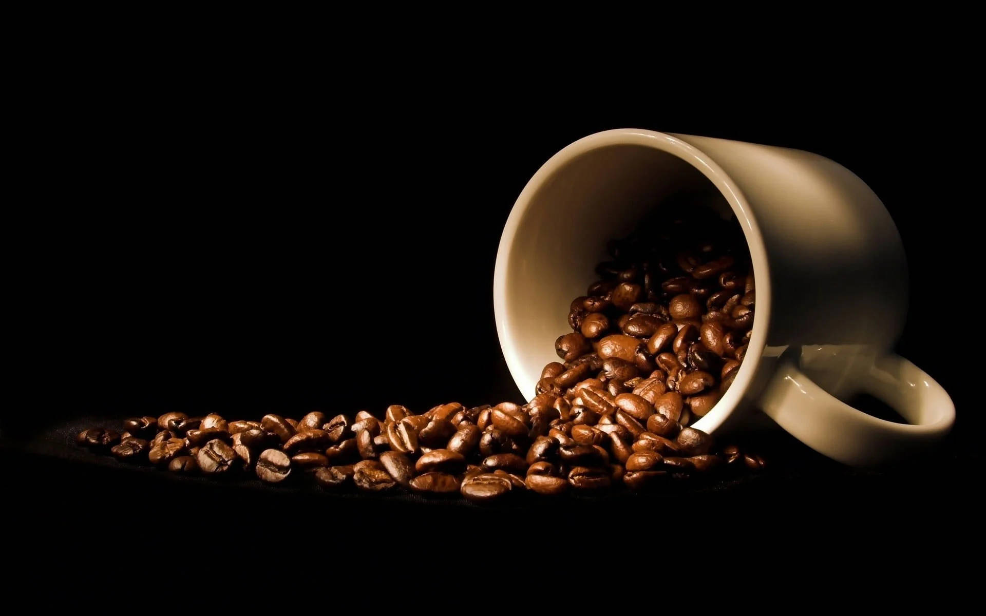 Coffee Aesthetic 桌布 壁紙 — 一個白色陶瓷杯在戲劇性燈光下將濃郁的棕色咖啡豆灑在黑色表面上。