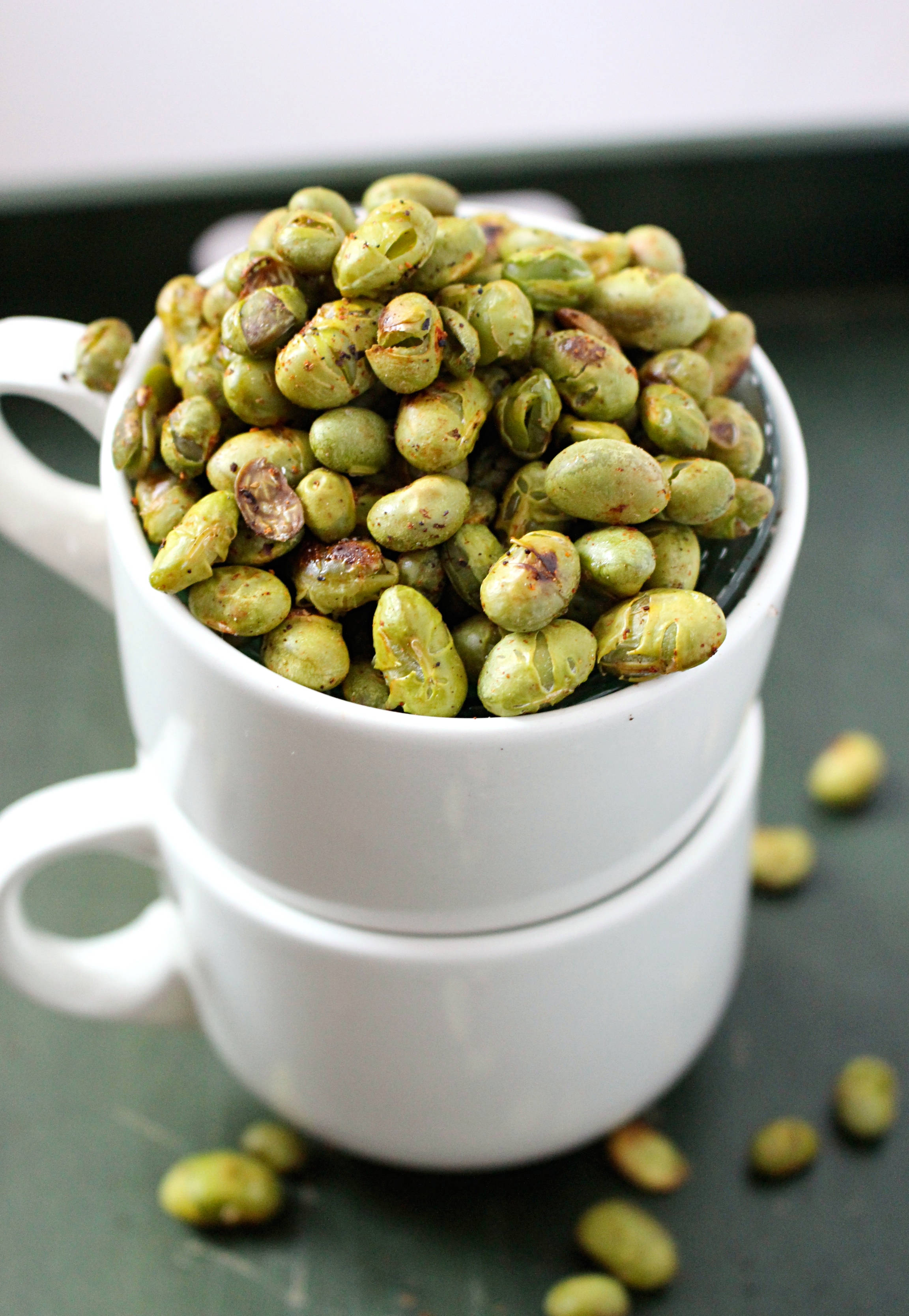 beans 桌布 壁紙 — 一個白色陶瓷杯疊在另一個杯子上，盛載著大量烤青枝豆，滿溢至杯邊，並有數顆散落在深色表面上。