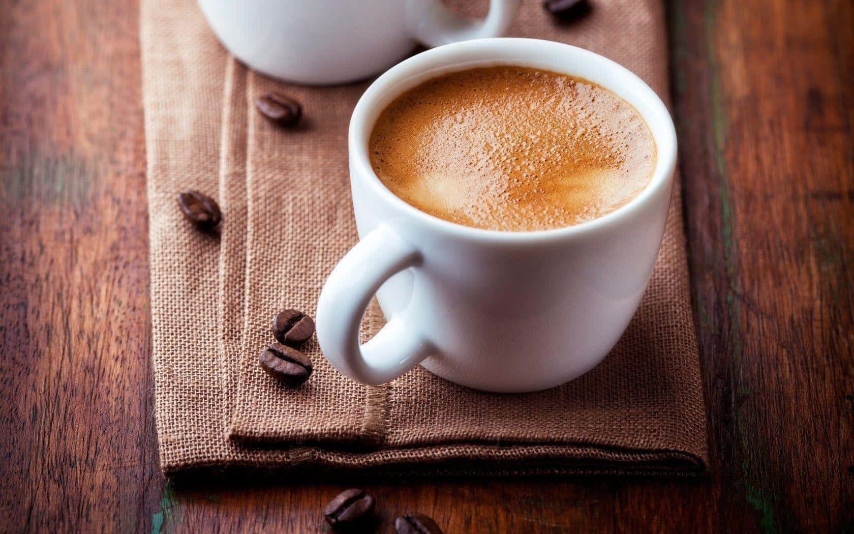 Coffee Aesthetic 桌布 壁紙 — 一個白色陶瓷杯裝滿泡沫咖啡，放在一個棕色粗麻布餐巾上，上面散落著烘焙咖啡豆，置於鄉村風木桌上。