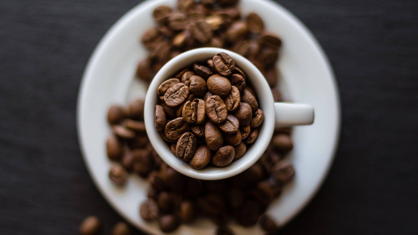 beans 桌布 壁紙 — 一個白色陶瓷杯裝滿烘焙咖啡豆，放在相配的碟子上，背景是深色有紋理的。