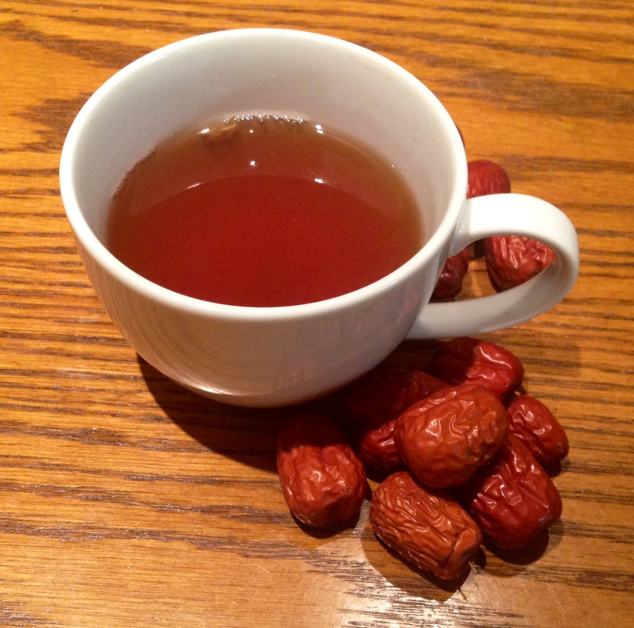 Fruits 桌布 壁紙 — 一個白色陶瓷杯裝滿琥珀色茶，放在溫暖的木桌上，旁邊是飽滿的紅棗。