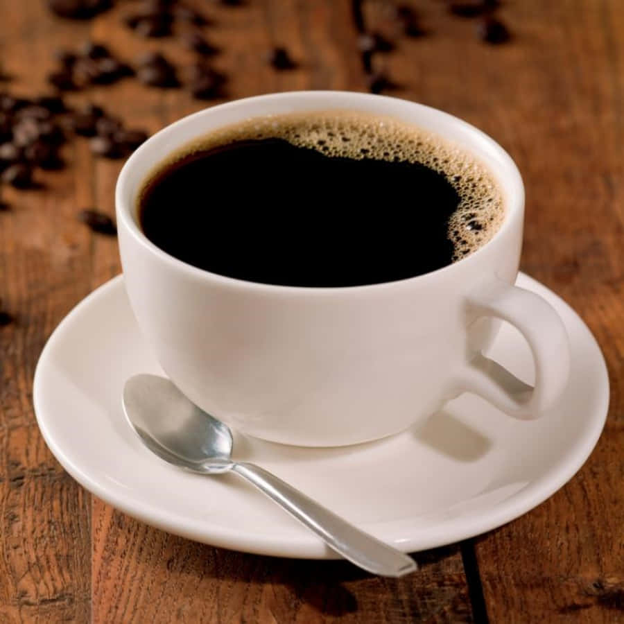 Coffee Cup 桌布 壁紙 — 一個白色陶瓷杯裝著黑咖啡和泡沫，放在碟子上，附有湯匙，置於散落咖啡豆的鄉村風格木桌上。