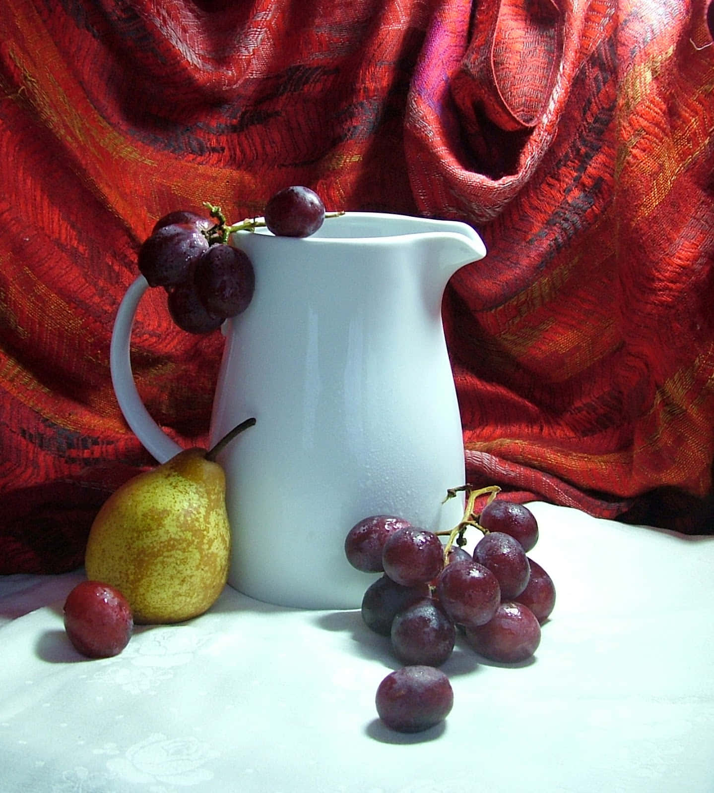 still life 桌布 壁紙 — 一個白色陶瓷水壺和新鮮葡萄放在一塊白布上，背景是豐富的紅色紋理布料。
