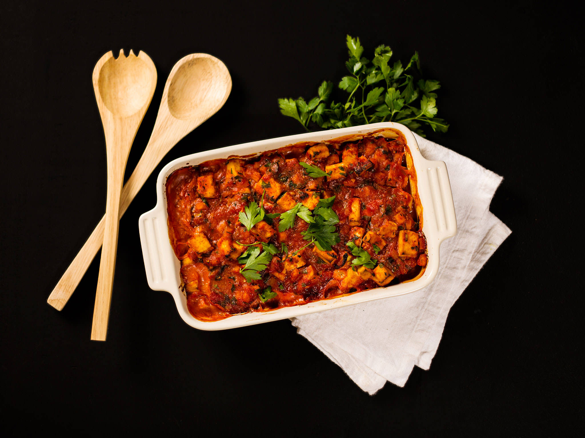 Food Photography 桌布 壁紙 — 一個白色陶瓷烤盤，盛滿濃郁的紅色番茄燉菜，上面撒上新鮮歐芹，放在白色布上，背景為黑色，附近有木勺和新鮮香草。