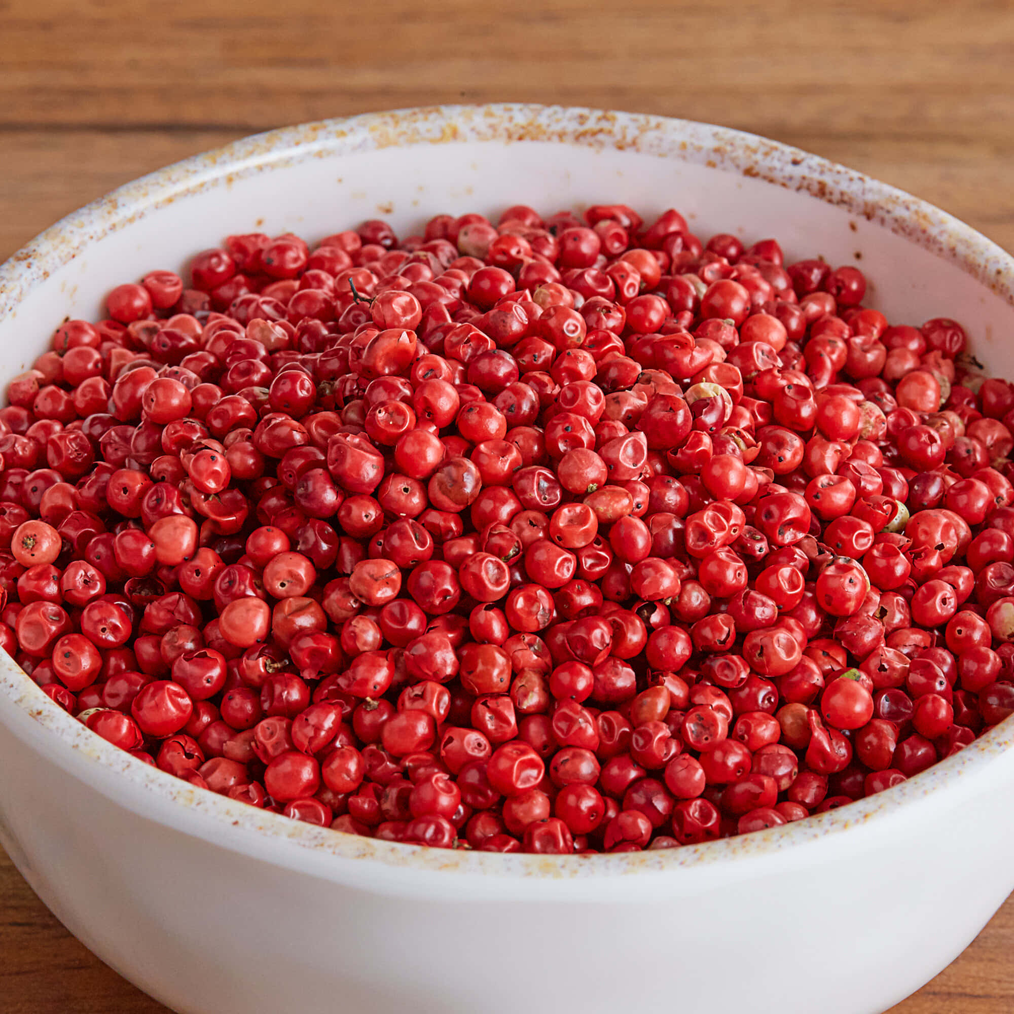 Pink Peppercorn 桌布 壁紙 — 一個白色陶瓷碗盛滿光澤的紅色胡椒粒，放在溫暖的木質表面上。
