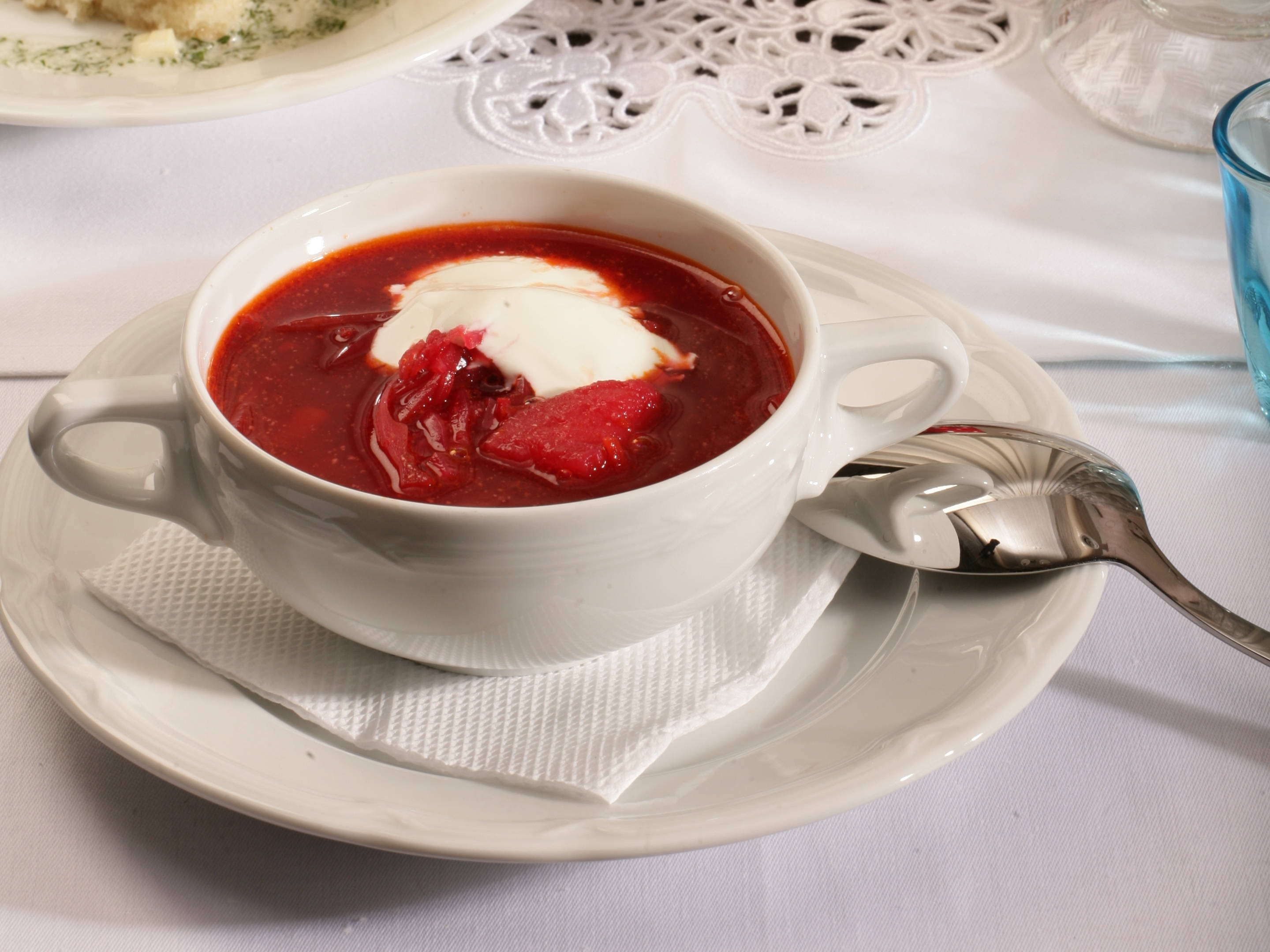 beetroot 桌布 壁紙 — 一個白色陶瓷碗盛滿濃郁的紅色羅宋湯，上面放有酸奶油和一勺甜菜根，放在蕾絲墊布上，旁邊有一隻湯匙。