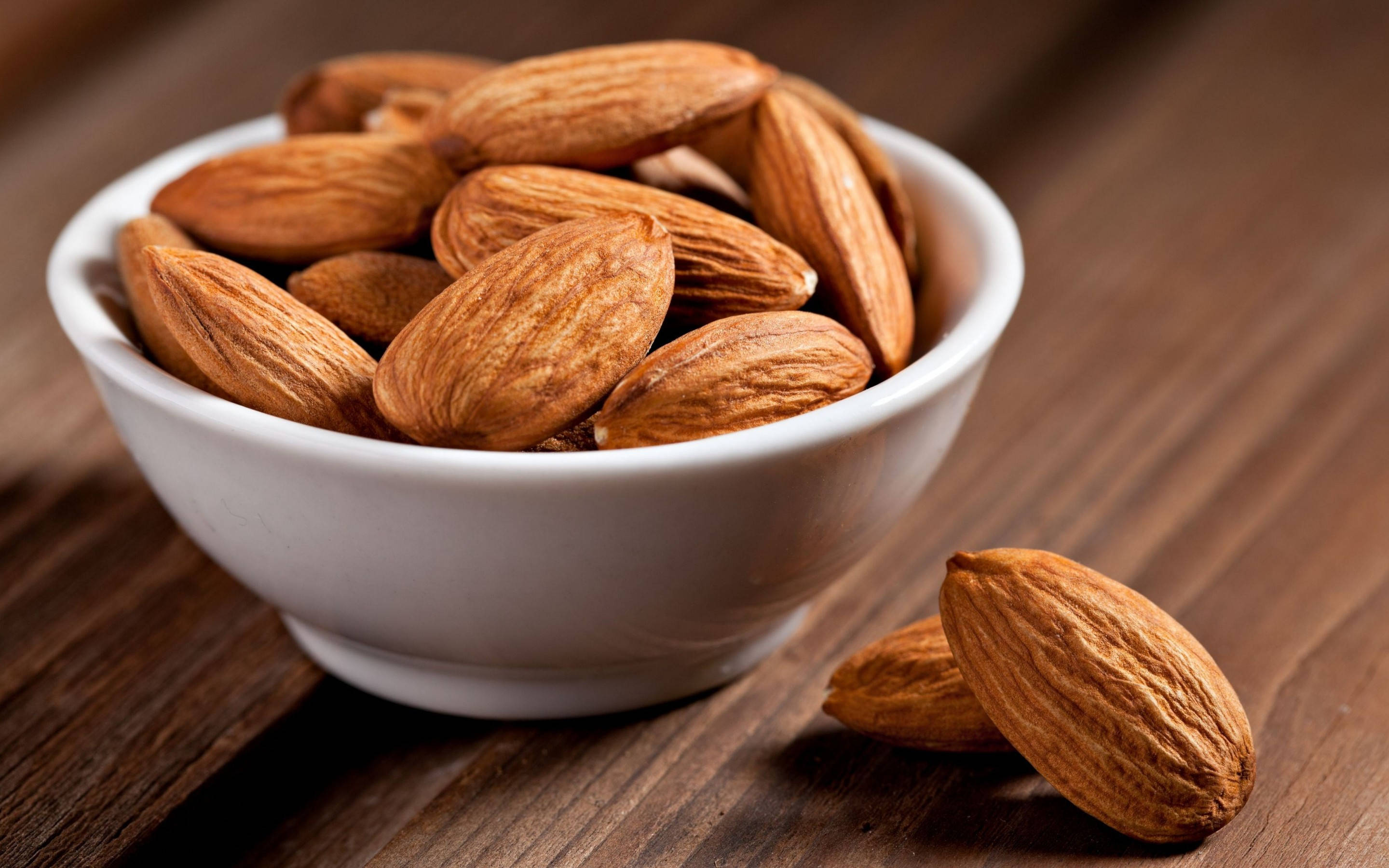 Almond 桌布 壁紙 — 一個白色陶瓷碗裝滿了質感豐富的棕色杏仁，放在深色木質表面上，附近有兩顆散落的杏仁。