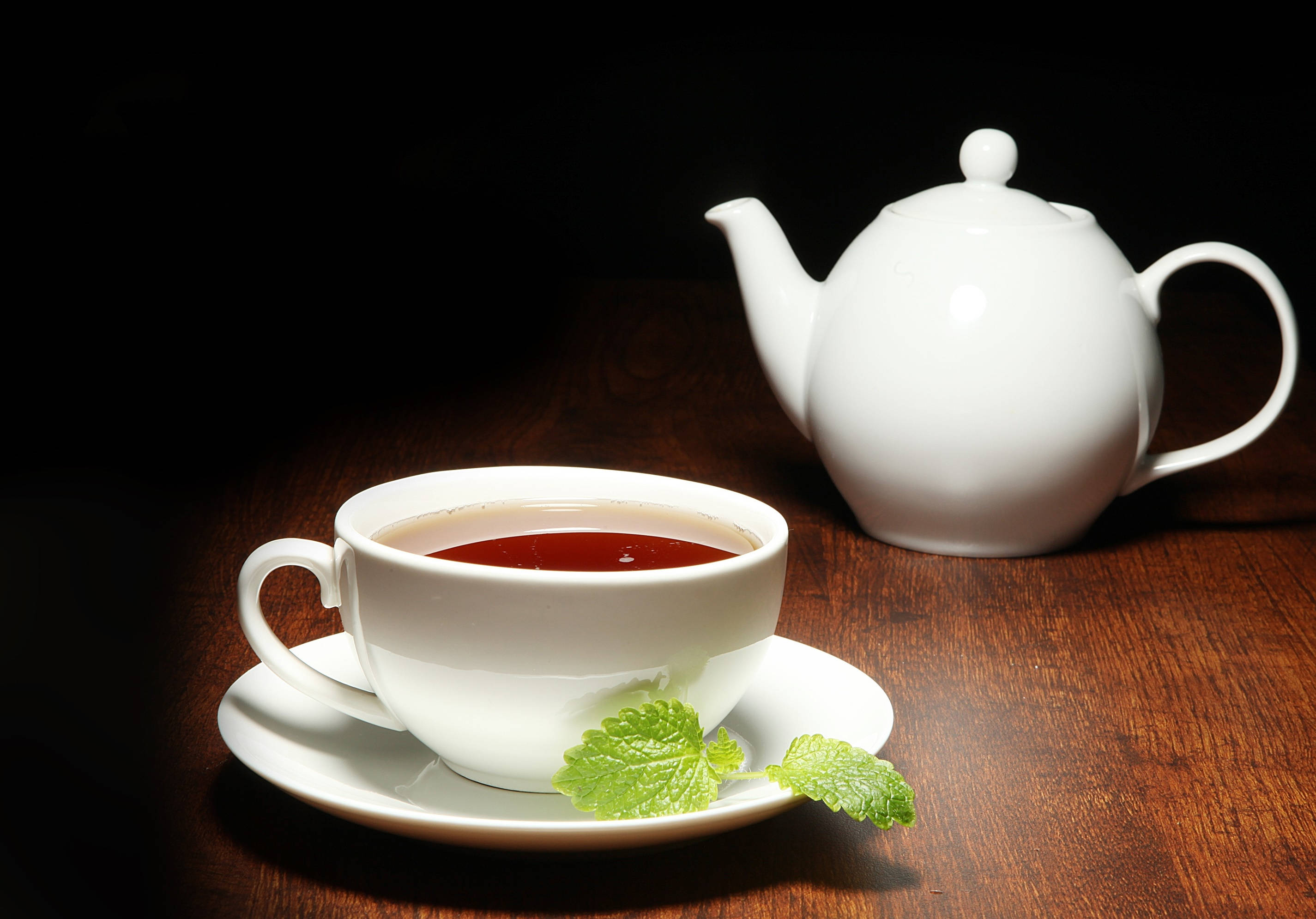 木紋 桌布 壁紙 — 一個白色陶瓷茶杯，裝滿琥珀色茶和新鮮薄荷葉，旁邊放著一個相配的茶壺，在深色木桌上，戲劇性的聚光燈下。