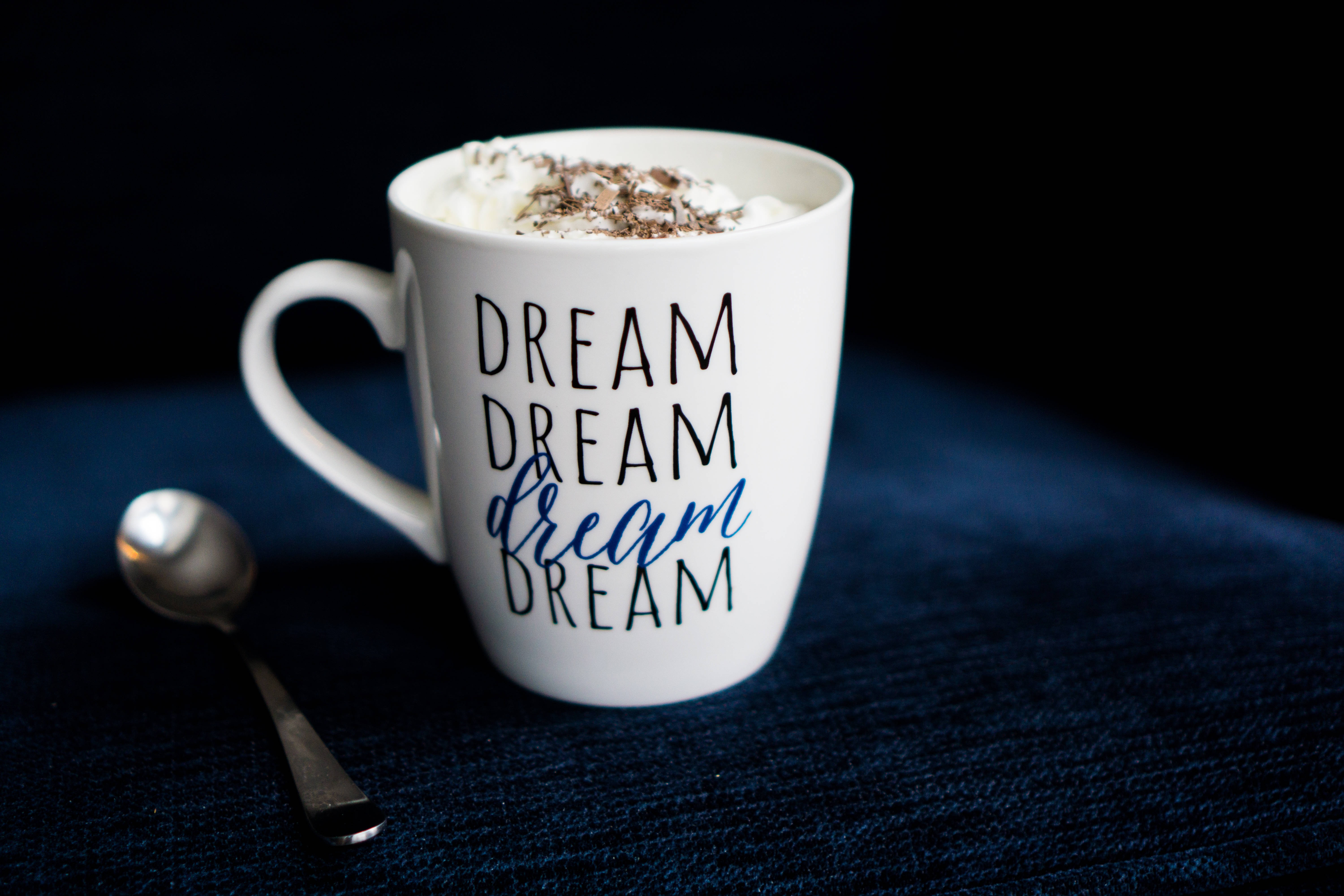 夢幻 桌布 壁紙 — 一個白色馬克杯，上面有「dream」字樣和鮮奶油，放在深藍色紋理表面上，旁邊有一隻湯匙。