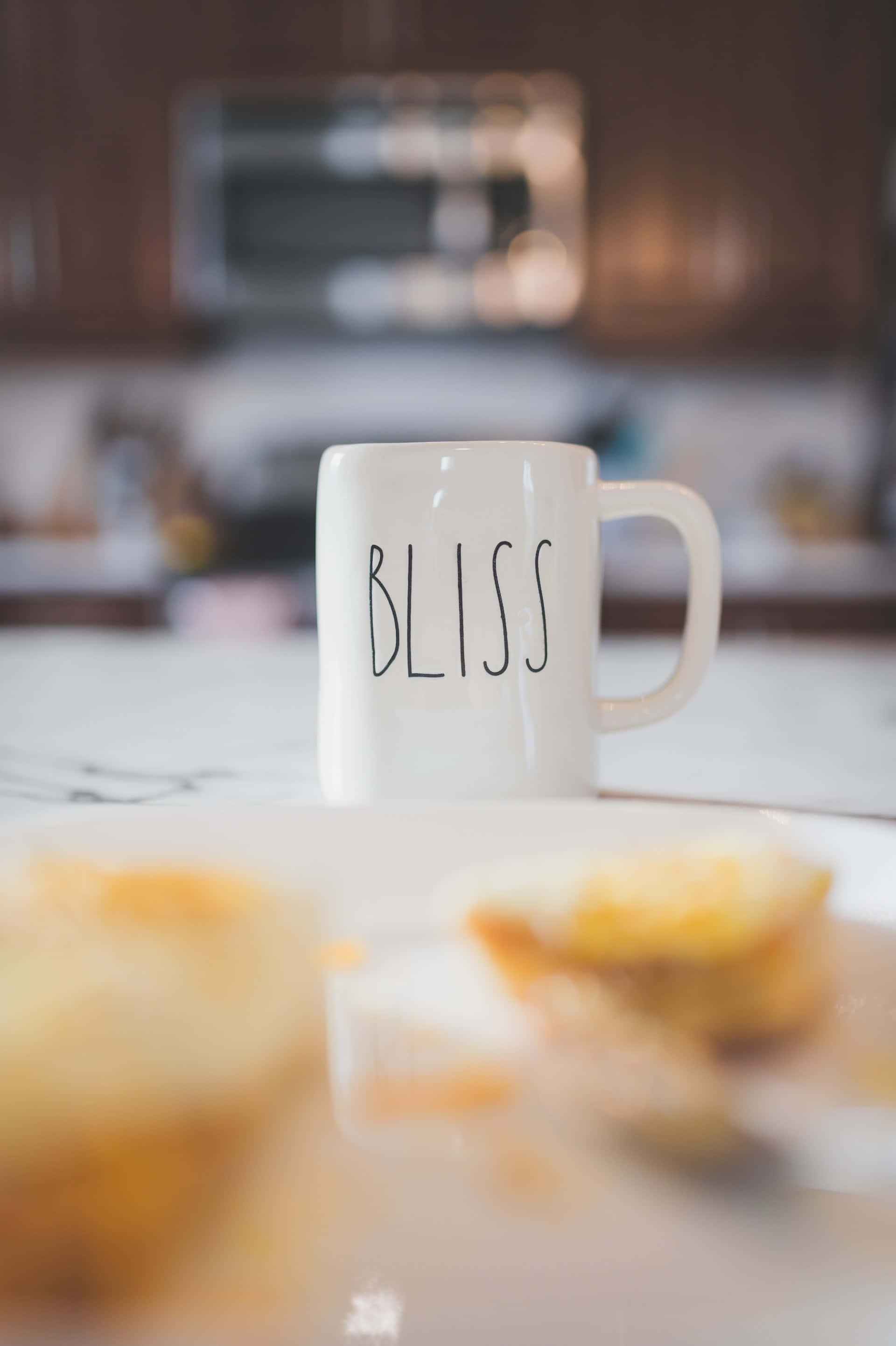 Blissful 桌布 壁紙 — 一個白色馬克杯，上面有黑色字體的 bliss 字樣，放在廚房檯面上，旁邊有一盤模糊的糕點。