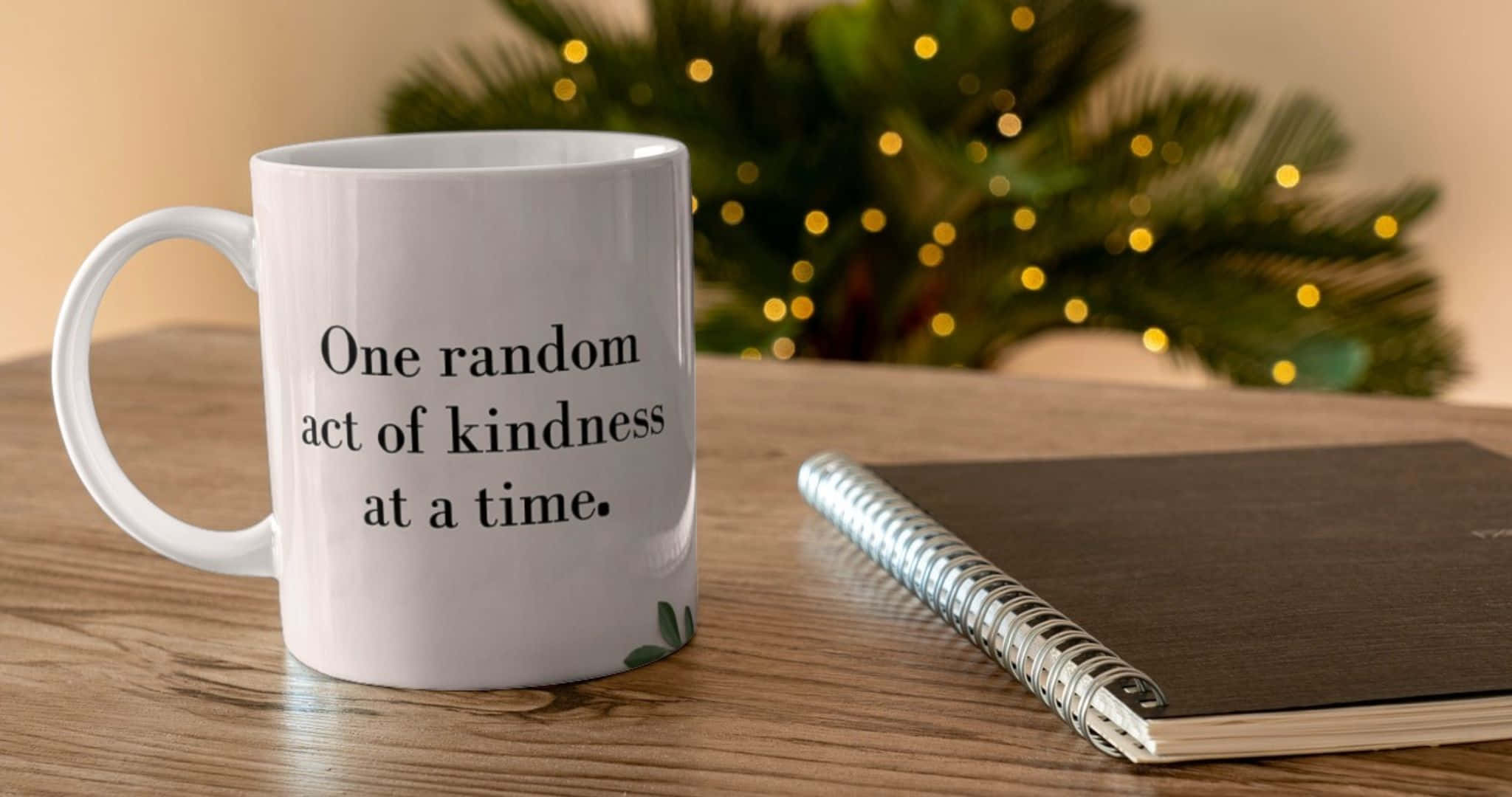 2048x1080 桌布 壁紙 — 一個白色馬克杯上寫著「one random act of kindness at a time」，旁邊放著一本筆記本，放在木桌上，背景是模糊的綠色植物和燈光。