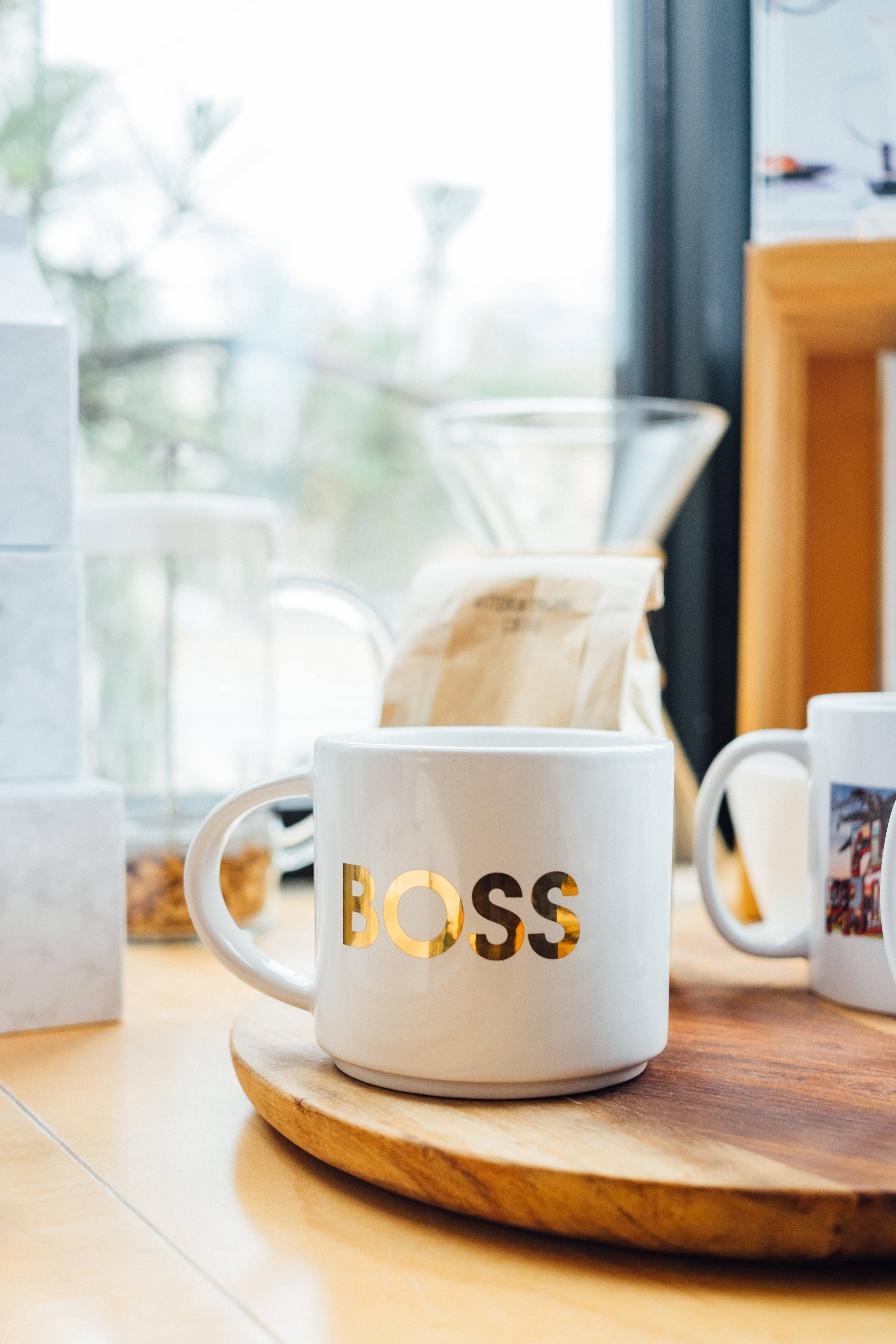 少女風 桌布 壁紙 — 一個白色馬克杯帶有金色 'BOSS' 字樣，放在木托盤上，旁邊有咖啡用具，靠近明亮的窗戶。