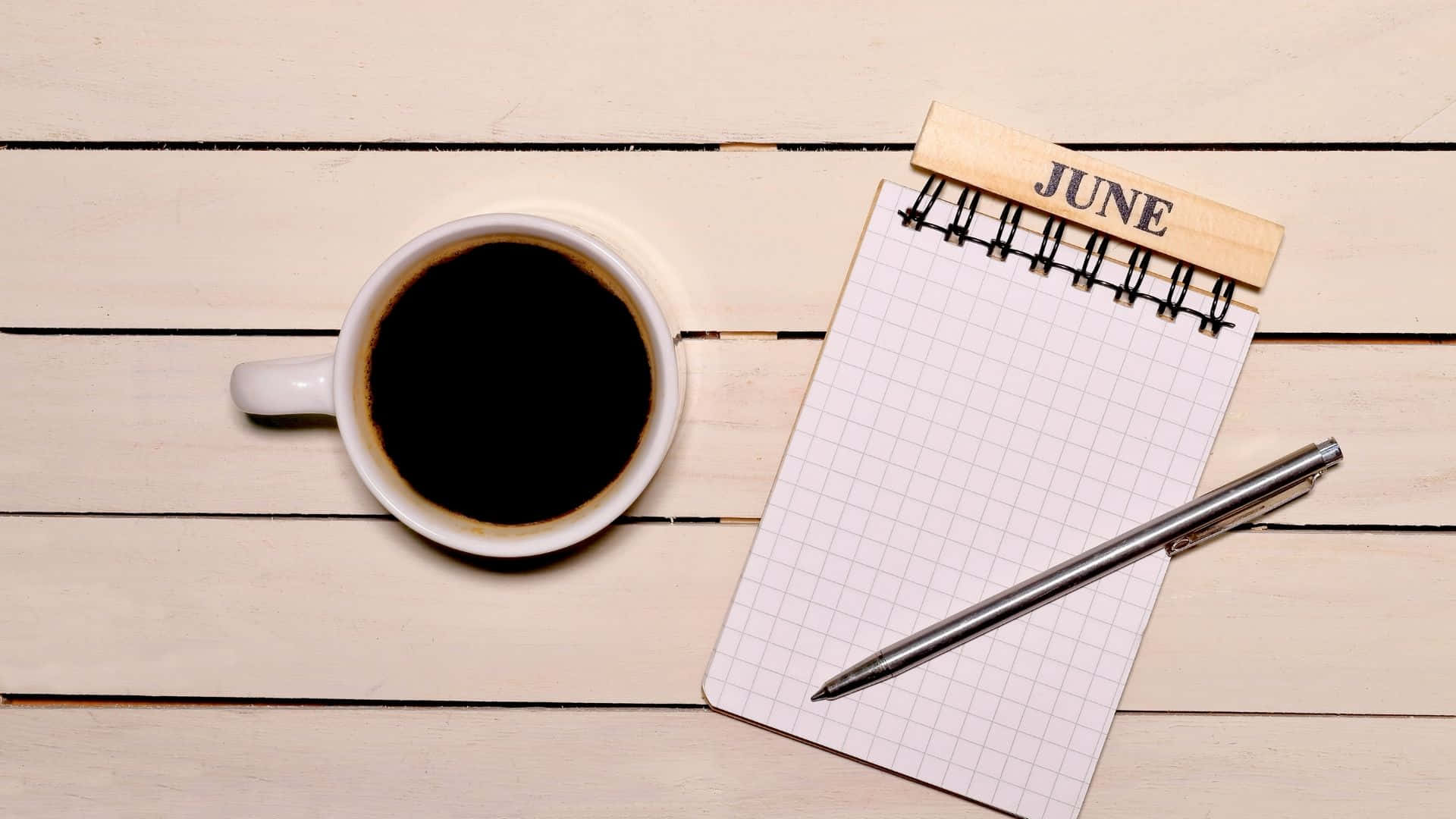 June 桌布 壁紙 — 一個白色馬克杯的黑咖啡放在淺色木板上，旁邊是一本六月主題的筆記本和筆。