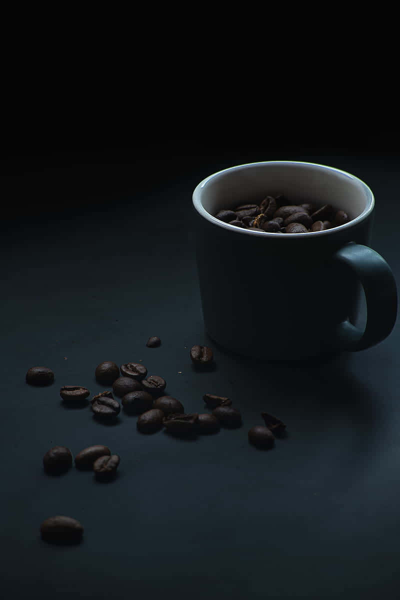 Coffee Aesthetic 桌布 壁紙 — 一個白色馬克杯裝滿深烘焙咖啡豆，溢出到黑色表面上，散落的豆子在戲劇性的低調燈光下。