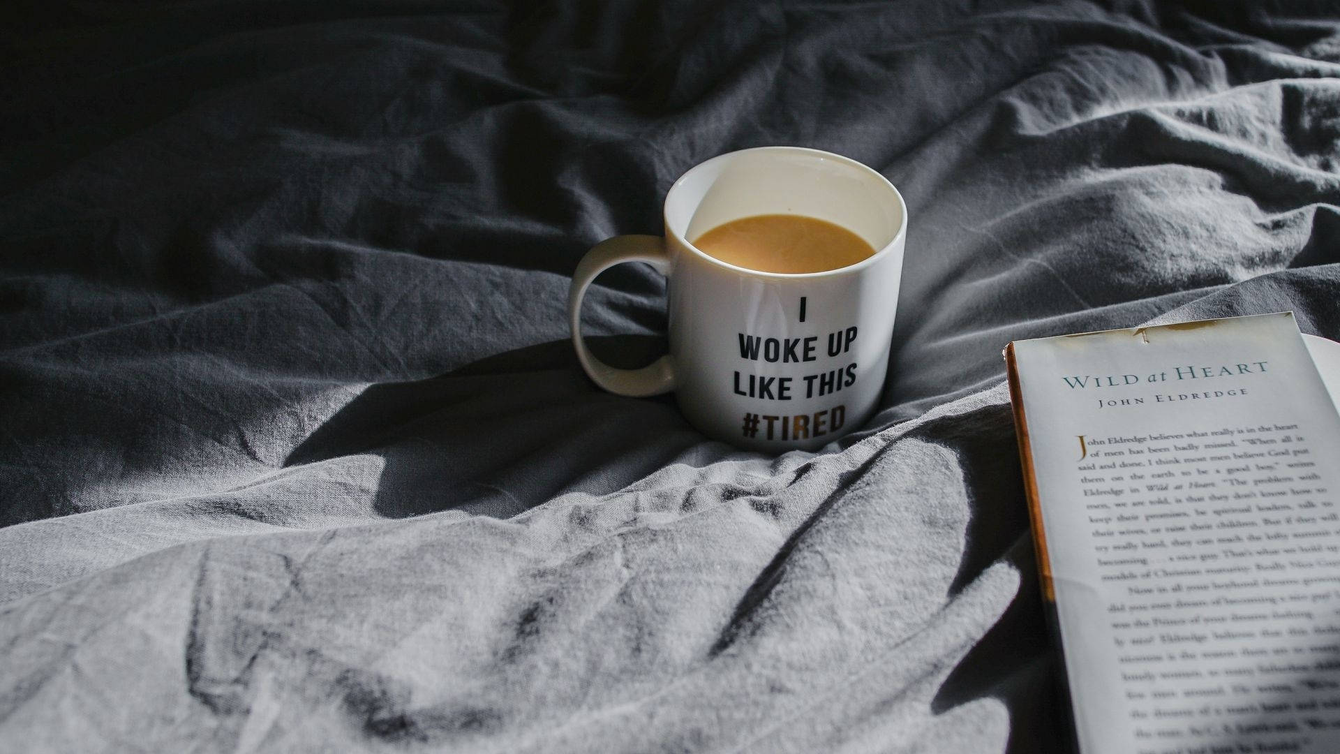 Coffee Aesthetic 桌布 壁紙 — 一個白色馬克杯裝著咖啡，上面有「I woke up like this #tired」的文字，旁邊放著一本打開的書，放在皺巴巴的灰色床單上。