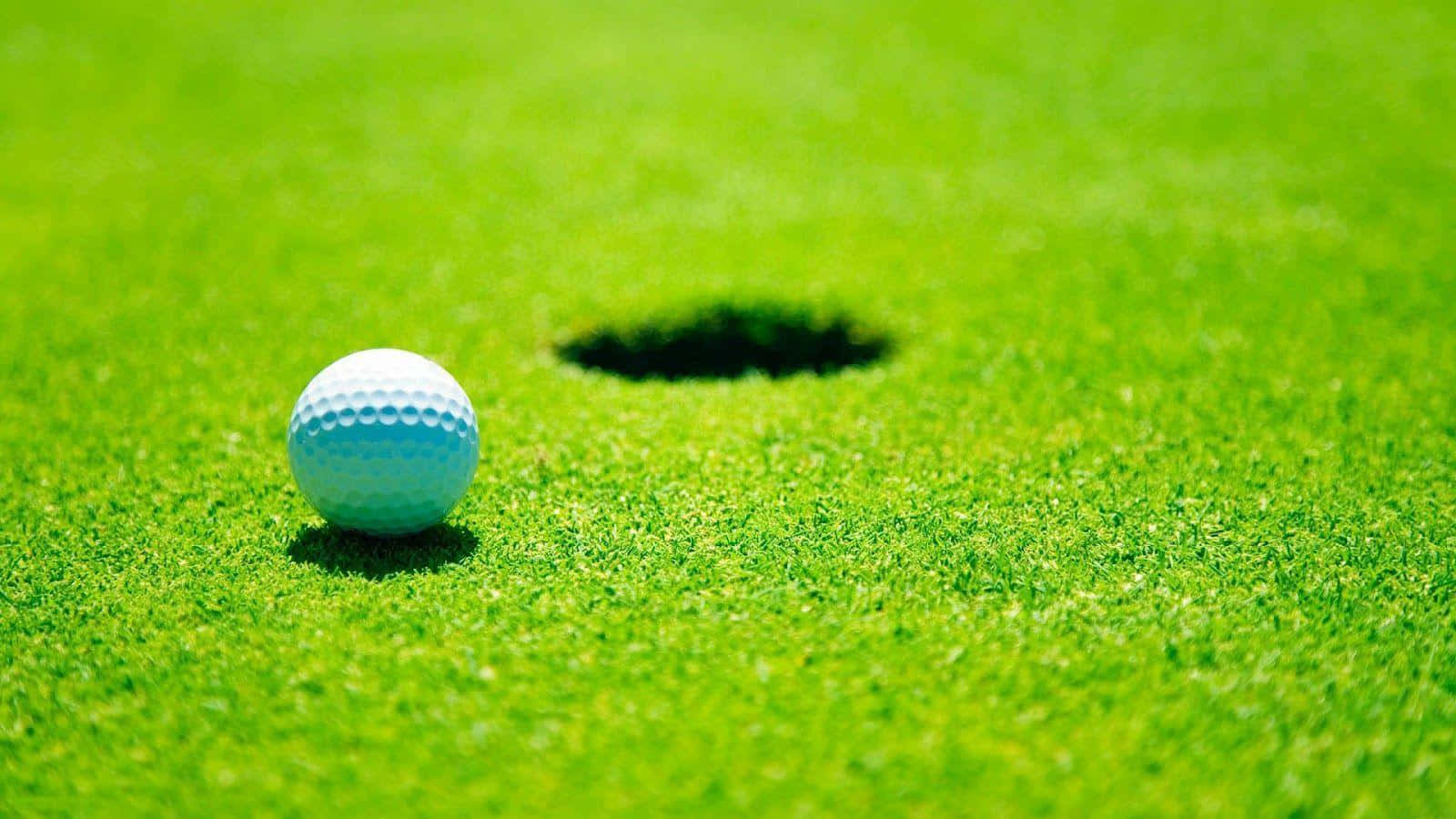sport 桌布 壁紙 — 一個白色高爾夫球在明亮的陽光下，停留在充滿活力的綠色草地上，旁邊是一個深色的球洞。