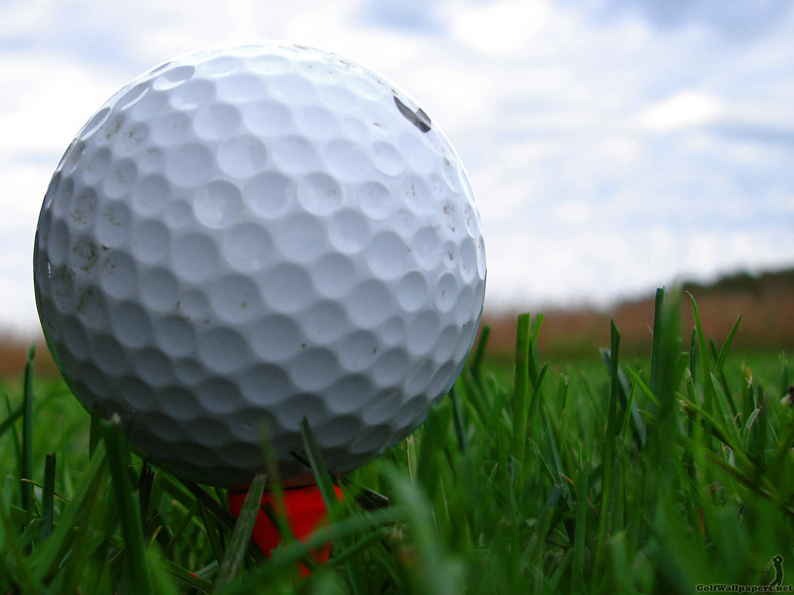 Golfing 桌布 壁紙 — 一個白色高爾夫球在紅色發球座上，置於充滿活力的綠色草地上，天空多雲的特寫。