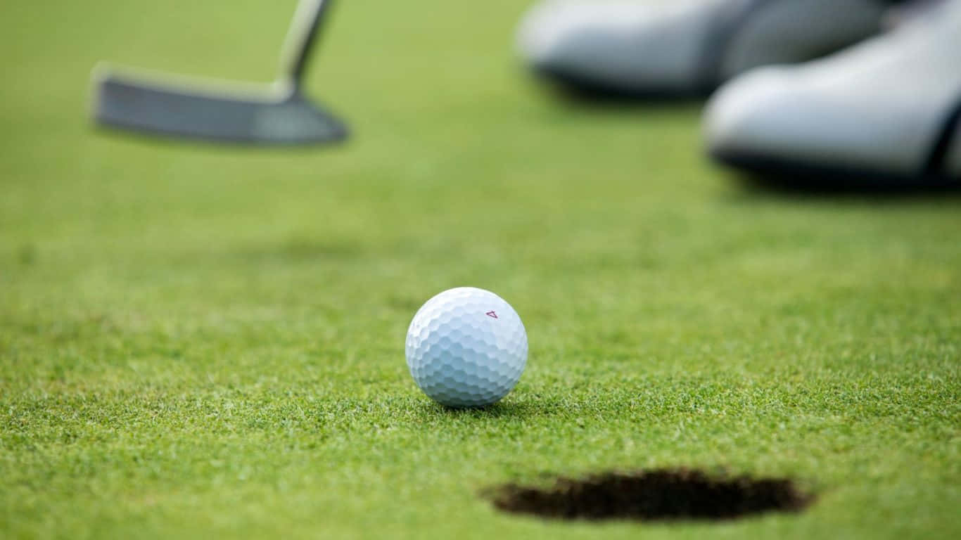 1366x768 Golf Course 桌布 壁紙 — 一個白色高爾夫球放在充滿活力的綠色草地上，靠近球洞，背景中有一個推桿和高爾夫球手的鞋子模糊不清。