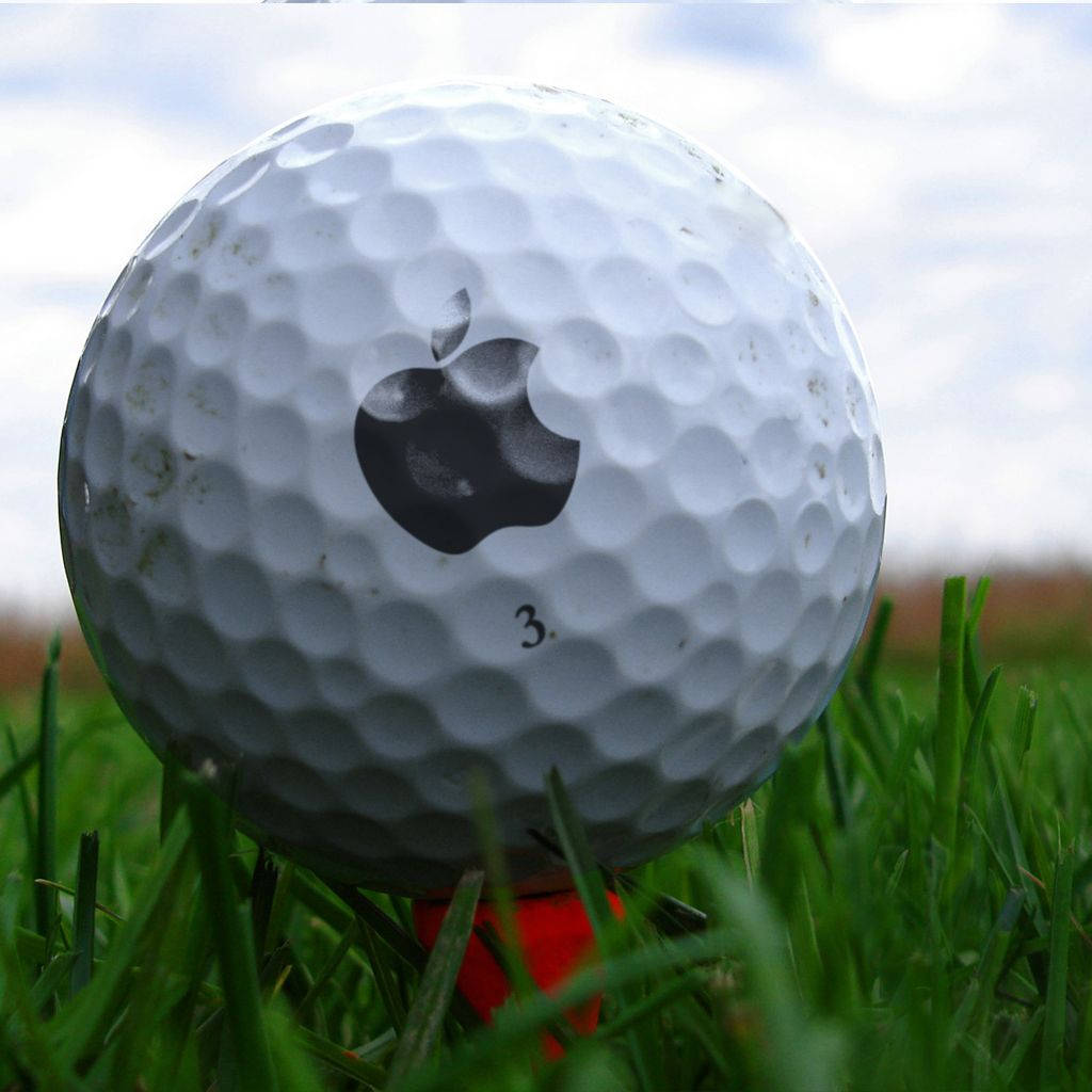 Golfing 桌布 壁紙 — 一個白色高爾夫球的特寫，帶有黑色Apple標誌，放在陰天天空下的綠色草地上。