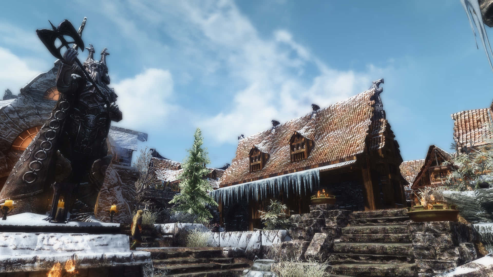 Skyrim Windhelm 桌布 壁紙 — 一個白雪皚皚的奇幻村莊，有一座高聳的盔甲雕像和掛滿冰柱的木屋，在明亮的藍天下。