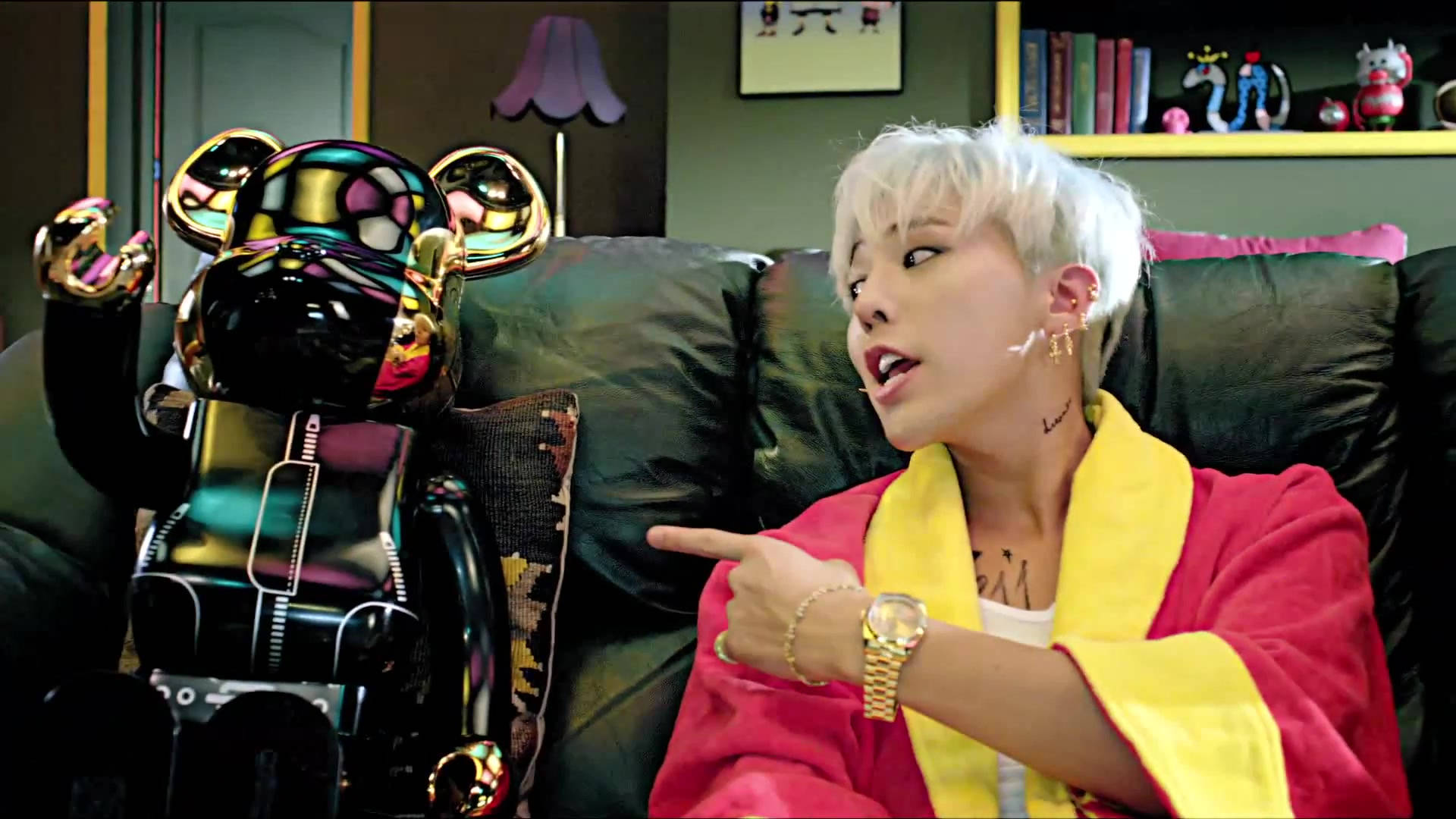 G-dragon 桌布 壁紙 — 一個白髮的人穿著紅色長袍，坐在深色沙發上指著一個閃亮的黑色熊雕像。
