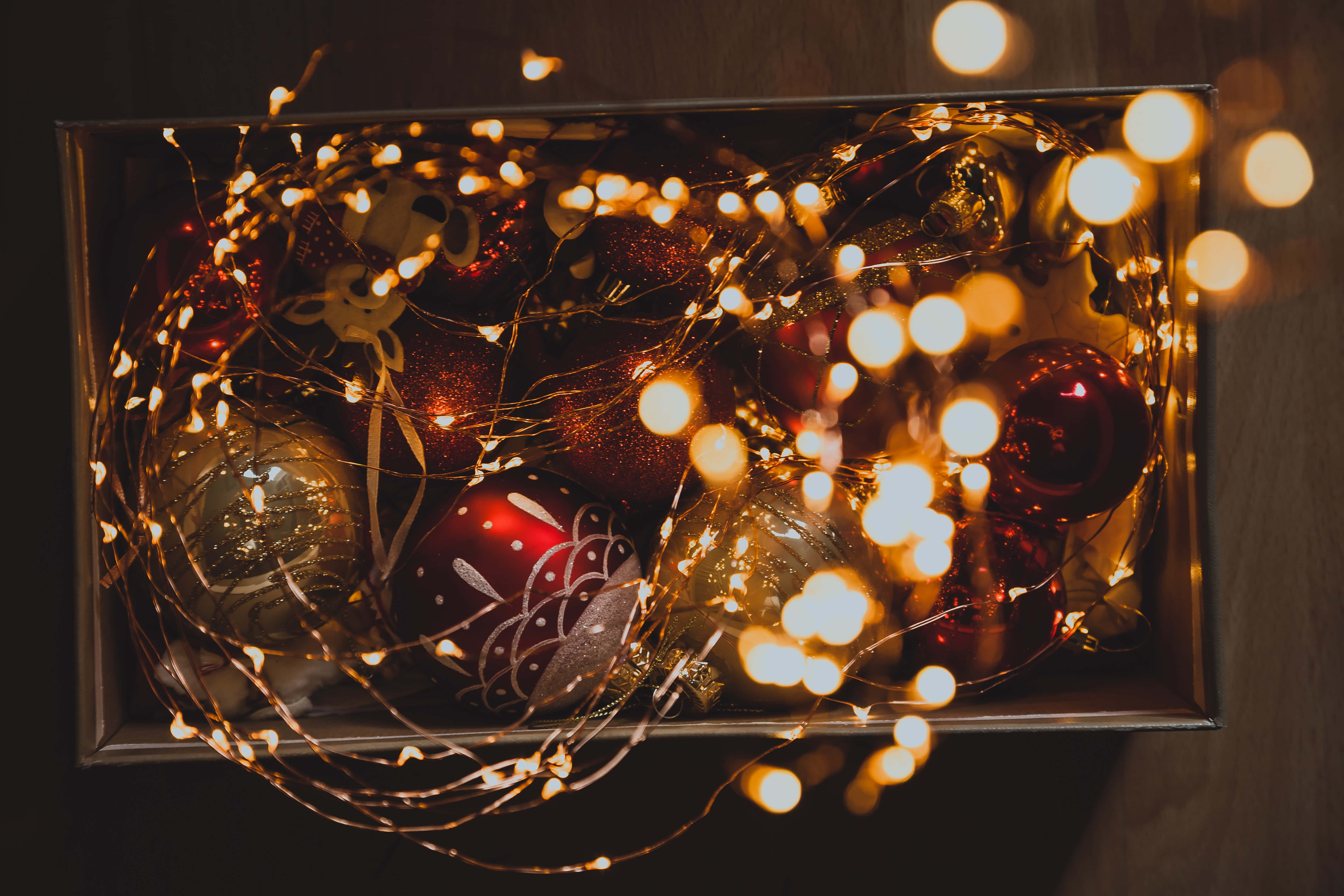 Christmas Lights Aesthetic 桌布 壁紙 — 一個盒子裡裝滿了節日紅色和金色的裝飾品，纏繞在發光的仙女燈中，背景是深色木質。
