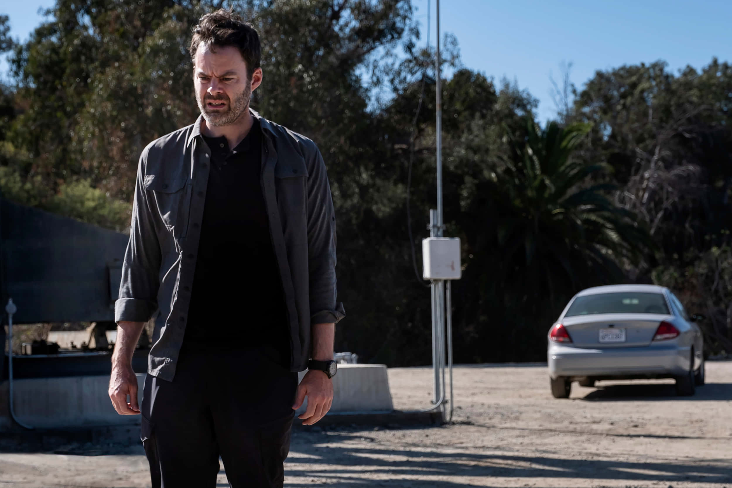 bill hader 桌布 壁紙 — 一個眉頭緊鎖、咬緊牙關的男人站在陽光普照的停車場，旁邊是一輛銀色汽車，背景有樹木。