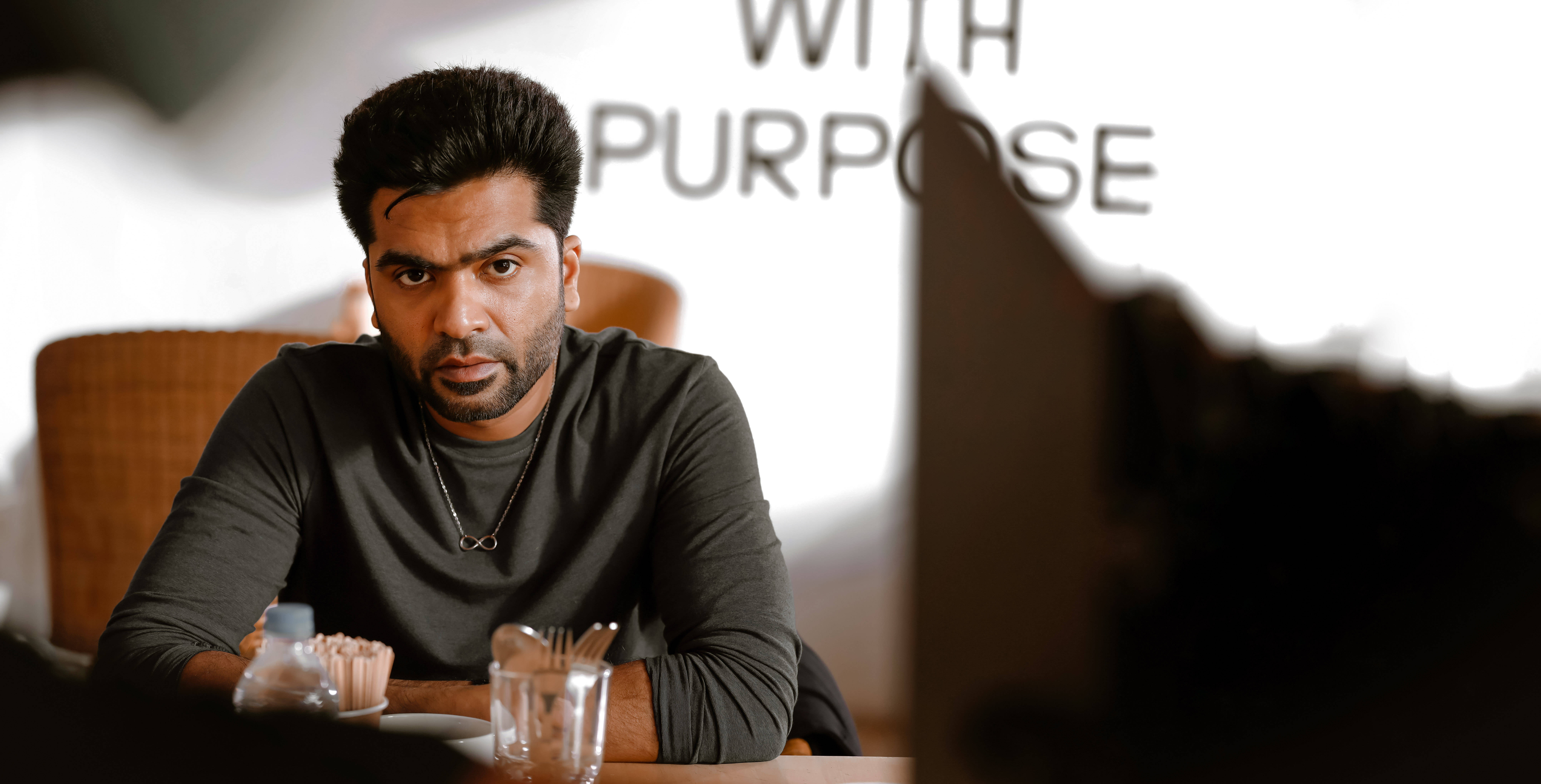 Indian 桌布 壁紙 — 一個眼神強烈的男人坐在咖啡店的桌子旁，身後有「WITH PURPOSE」的文字。