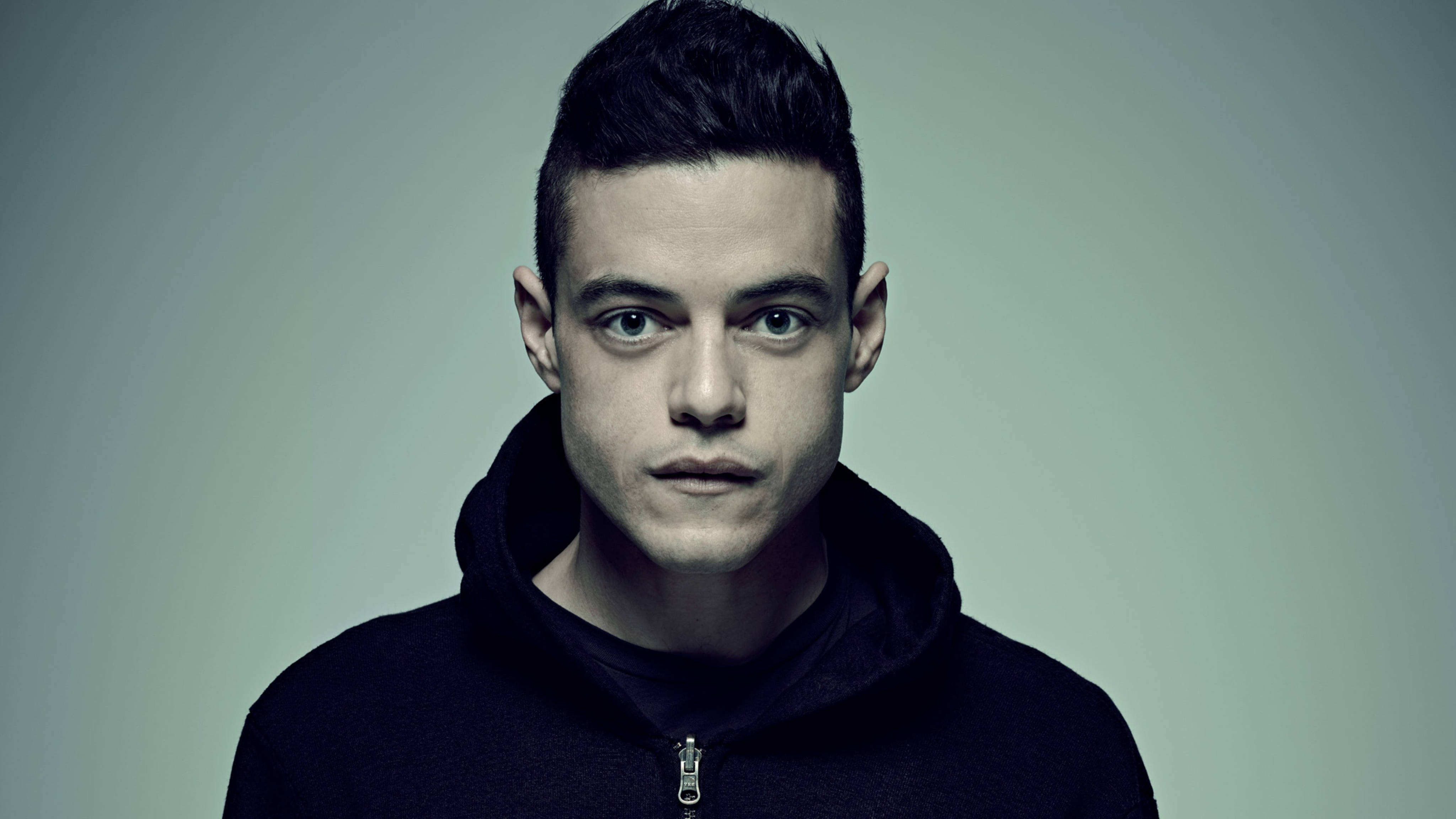 Mr Robot 桌布 壁紙 — 一個眼神銳利的年輕男子直接凝視鏡頭，身穿黑色連帽衫，背景為柔和的灰色。