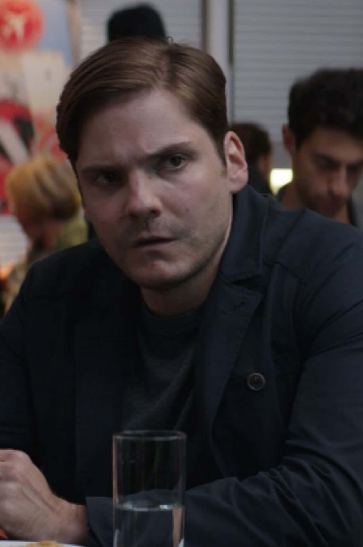Baron Zemo 桌布 壁紙 — 一個眼神銳利的男人坐在繁忙咖啡廳的桌子旁，穿著深色外套，直視鏡頭。