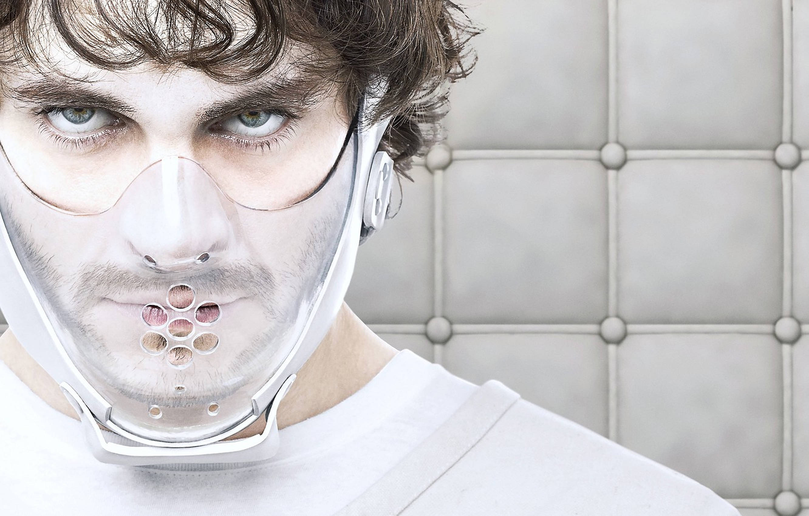 Hugh Dancy 桌布 壁紙 — 一個眼神銳利的男人戴著透明醫療口罩，帶有穿孔吹嘴，背景是簇絨灰色牆壁。