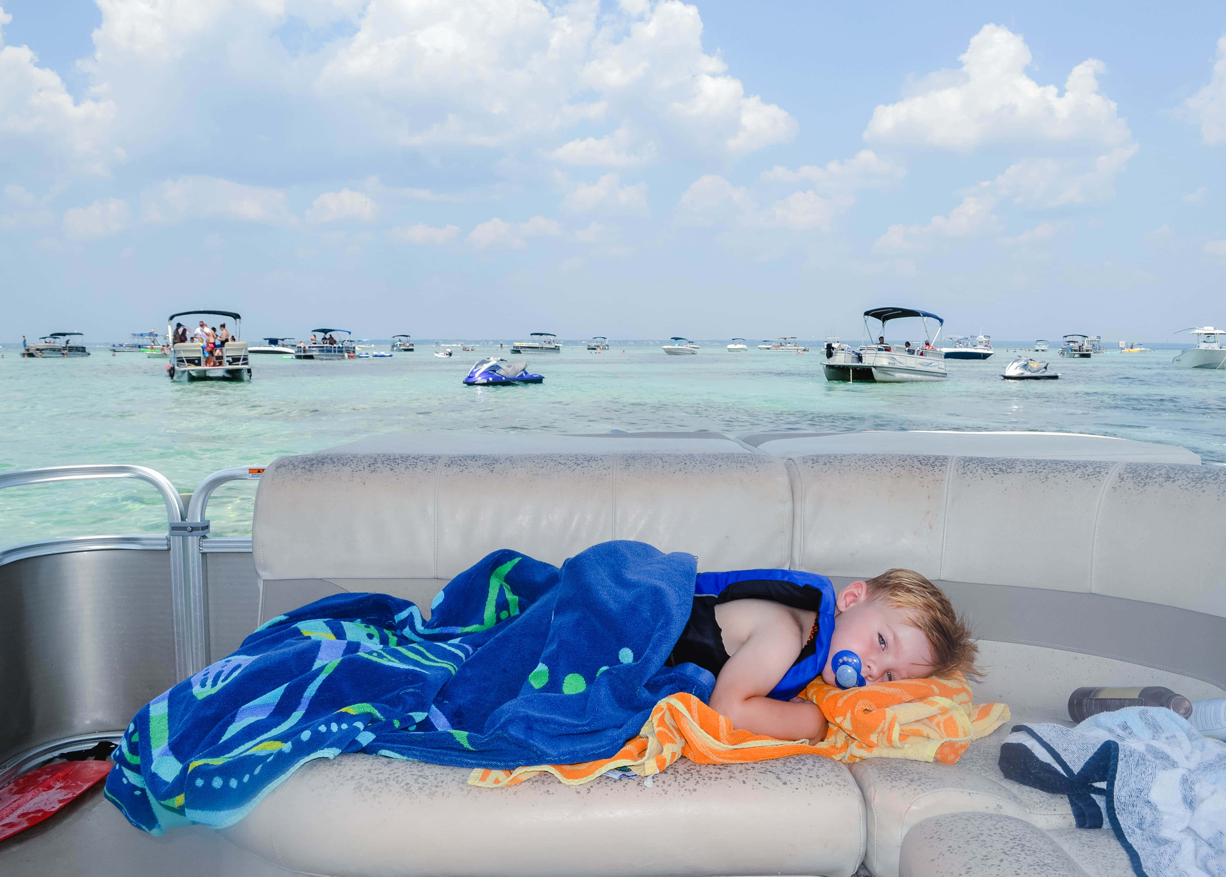 Crab Island 桌布 壁紙 — 一個睡眼惺忪的幼兒裹著藍色毛巾，在船墊上熟睡，其他船隻點綴在明亮的天空下的碧綠海水上。